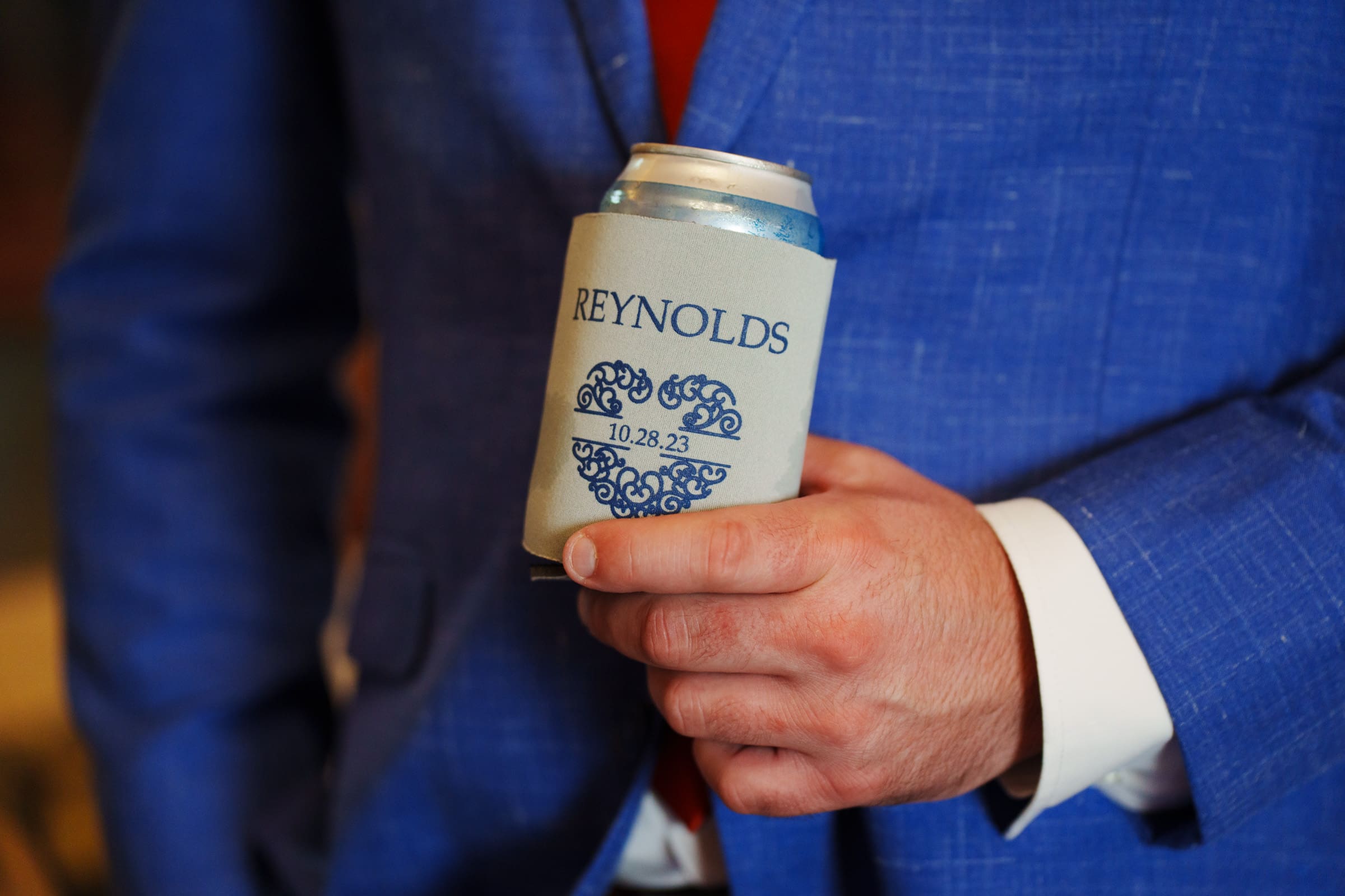 A person in a blue suit holds a beverage can wrapped in a custom koozie at the Rose Hill Estate wedding. The koozie is light gray with a blue floral heart design and the name Reynolds above a date: 10.28.23.