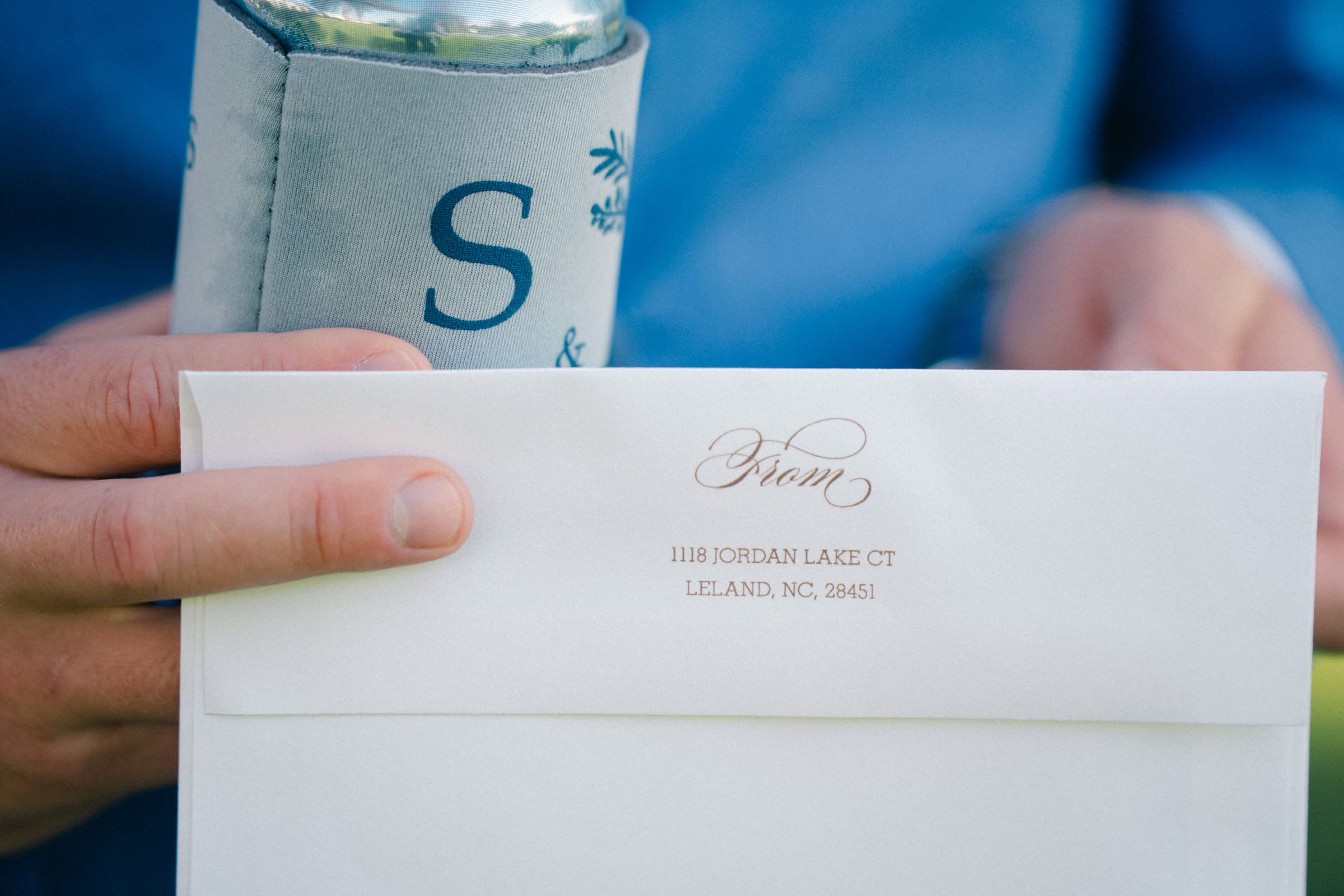 Close-up of hands holding a white envelope, elegantly scripted with an address: 1118 Jordan Lake Ct, Leland, NC, 28451. Nearby, a drink in a holder hints at the elegance of a Rose Hill Estate wedding as part of the letter S is visible.