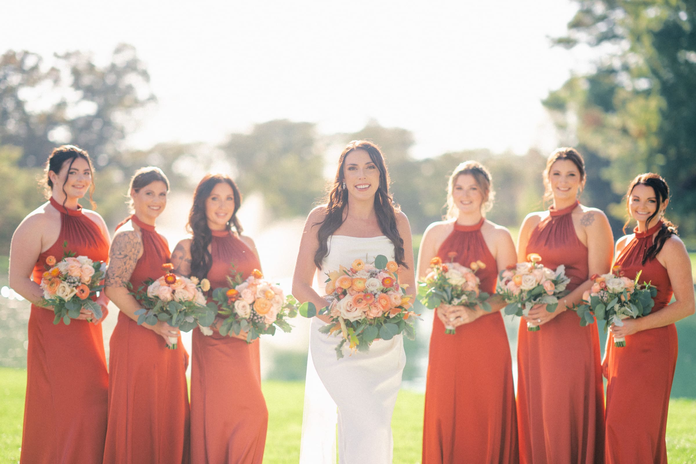 A bride stands outdoors at Rose Hill Estate with bridesmaids in matching orange dresses, each holding bouquets of flowers. The background reveals a grassy area with trees and a fountain in soft focus. The sun shines brightly, casting a warm, glowing effect over the charming wedding scene.