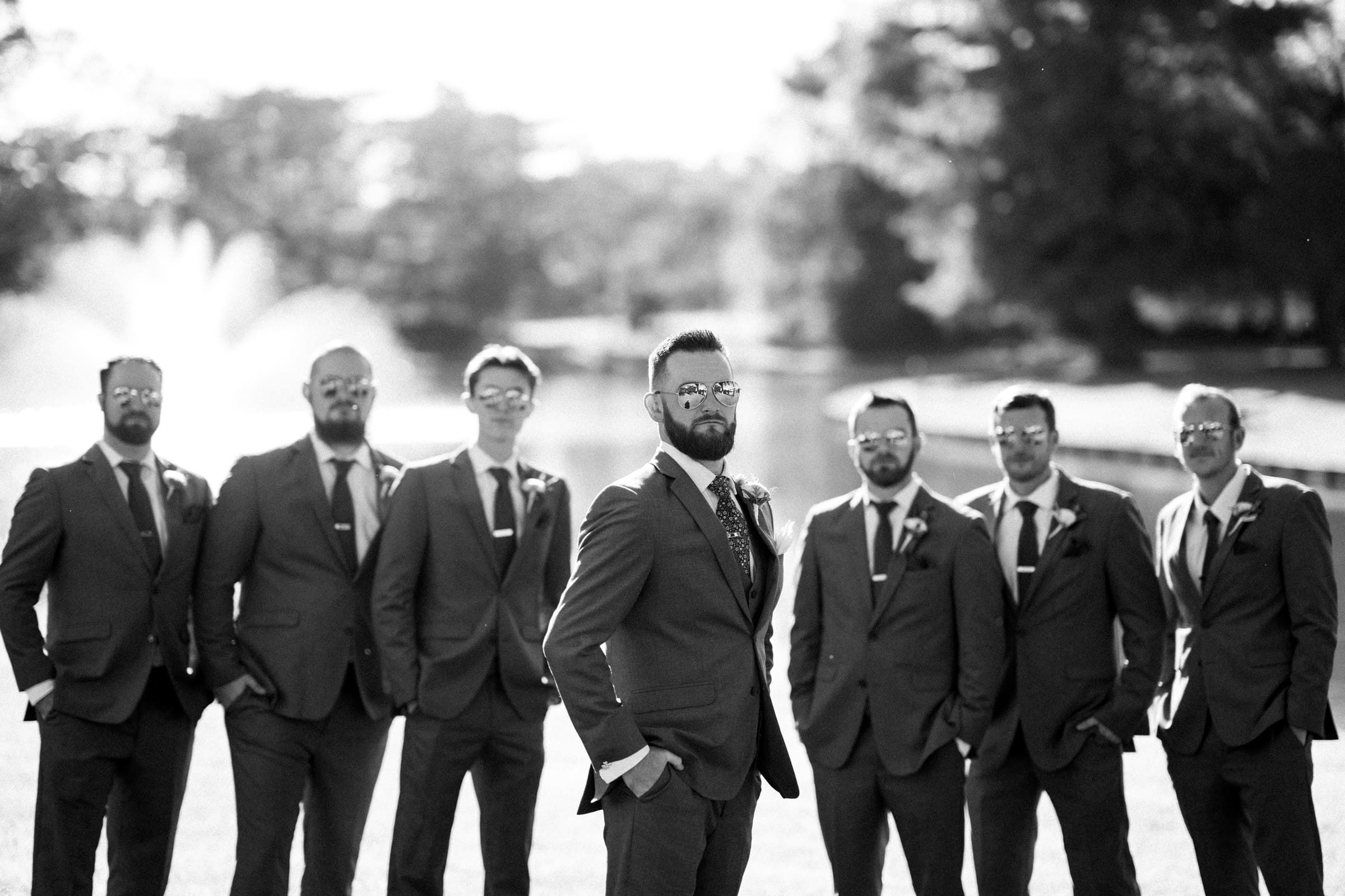 A black and white photo captures a group of seven men in suits and ties at a Rose Hill Estate wedding, standing outdoors. The man in the center stands slightly forward with his hands in his pockets, while trees and a fountain blur serenely in the background.