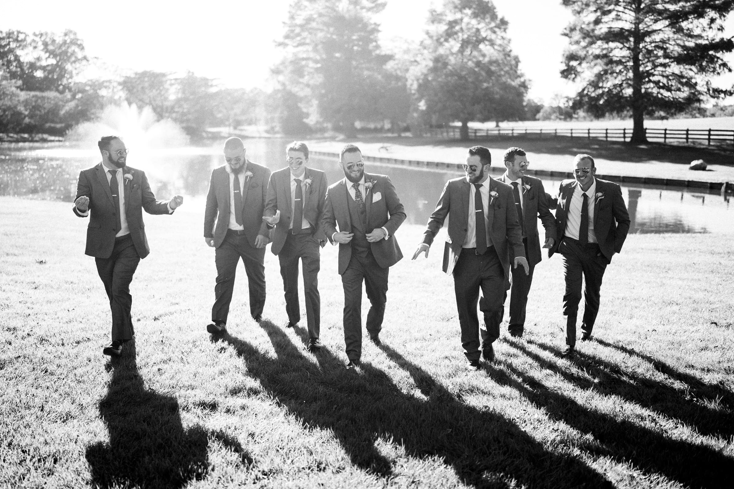 A group of seven men in suits walk happily across the grassy expanse of Rose Hill Estate, with trees and a pond in the background. The black-and-white scene captures their long shadows elegantly cast on the ground, adding a timeless charm to this wedding moment.