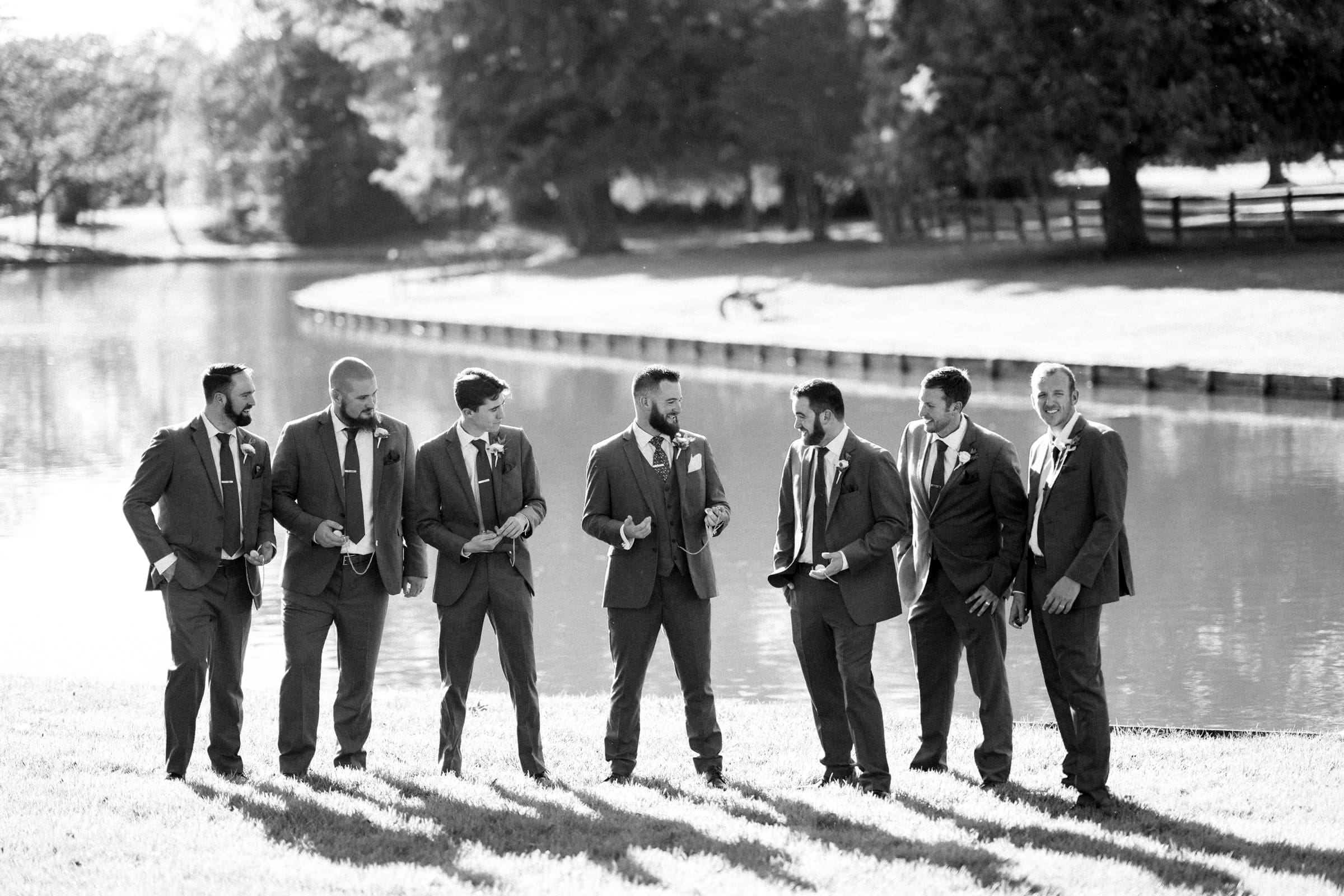 Seven men in suits stand in a grassy area near a pond at the Rose Hill Estate wedding, laughing and gesturing, with trees in the background. The scene is in black and white, capturing a lively moment.