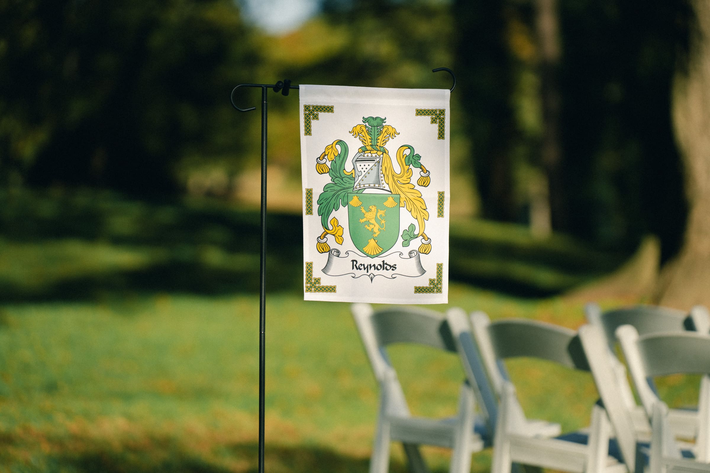 A small flag with a green and yellow coat of arms, featuring two lions and ornate details, graces a stand beside a row of white chairs in the lush outdoor setting of Rose Hill Estate, perfectly embodying the wedding's elegant ambiance.