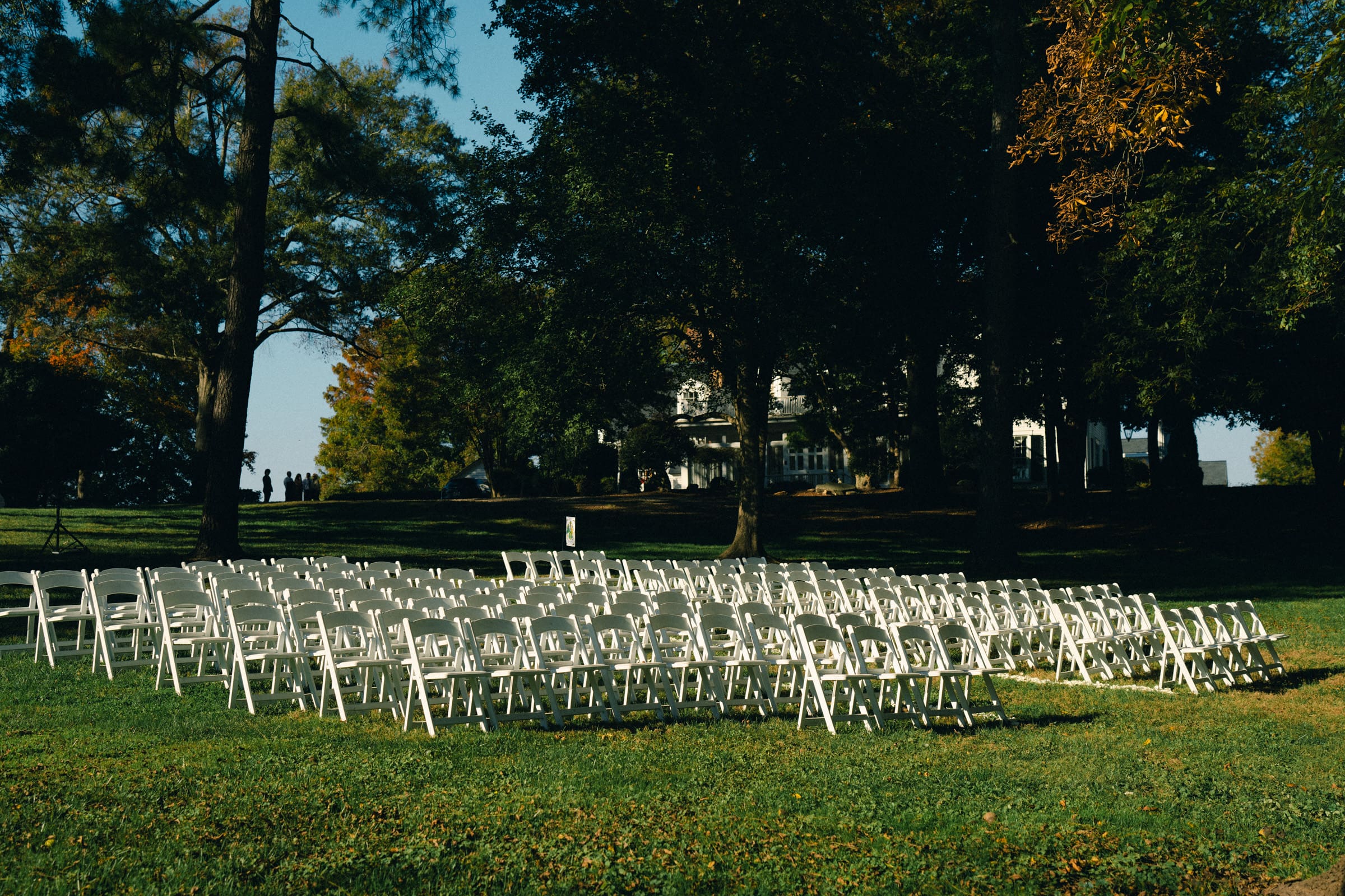 Rows of white folding chairs set up outdoors on a lush green lawn under tall trees, with the grand Rose Hill Estate visible in the background. The chairs are empty, suggesting preparations for a wedding event.
