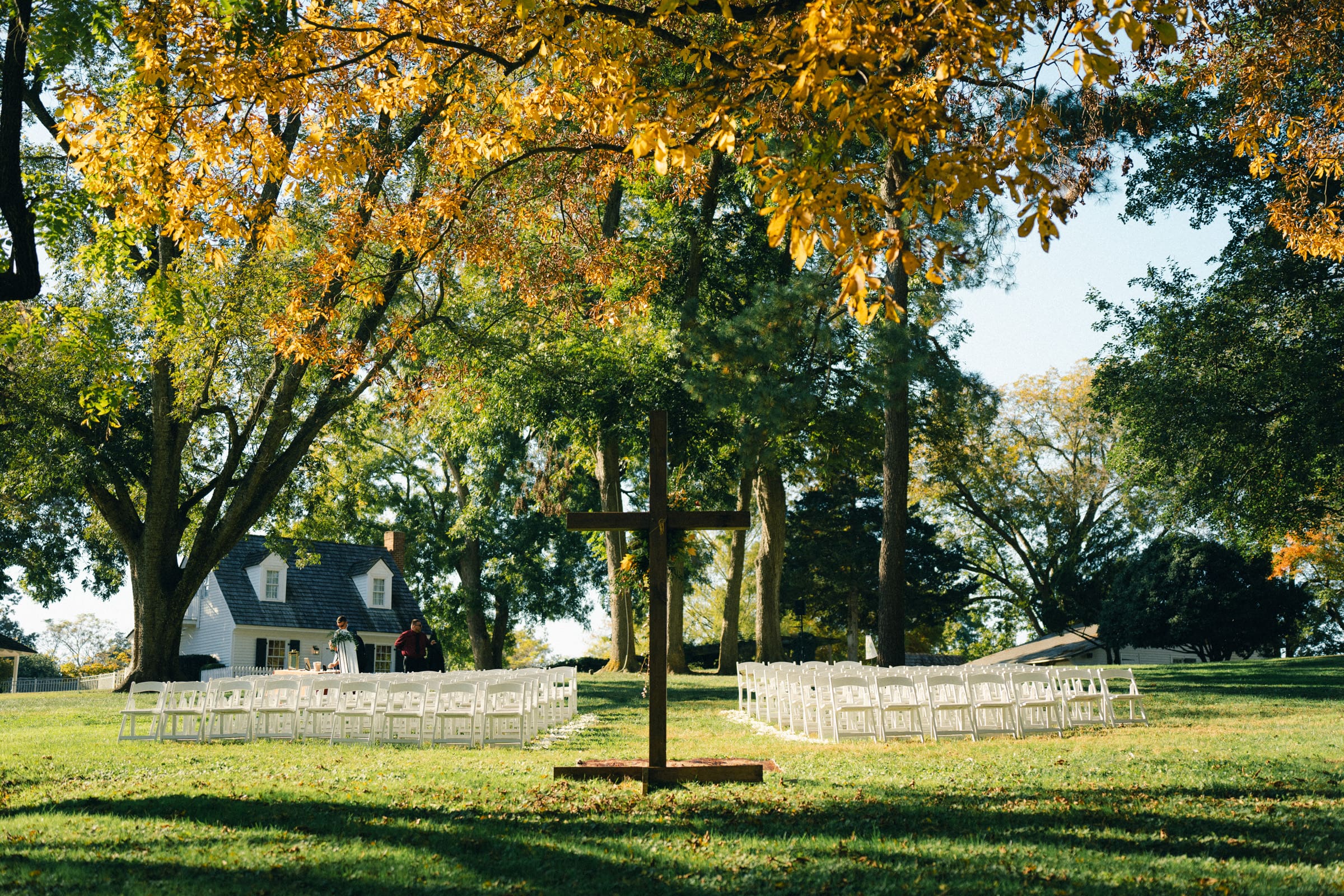 The serene outdoor setting at Rose Hill Estate features white chairs arranged in rows facing a large wooden cross on a lush green lawn. Surrounded by trees with autumn leaves, and with a quaint house in the background, this sunlit scene is perfect for a tranquil wedding ceremony.