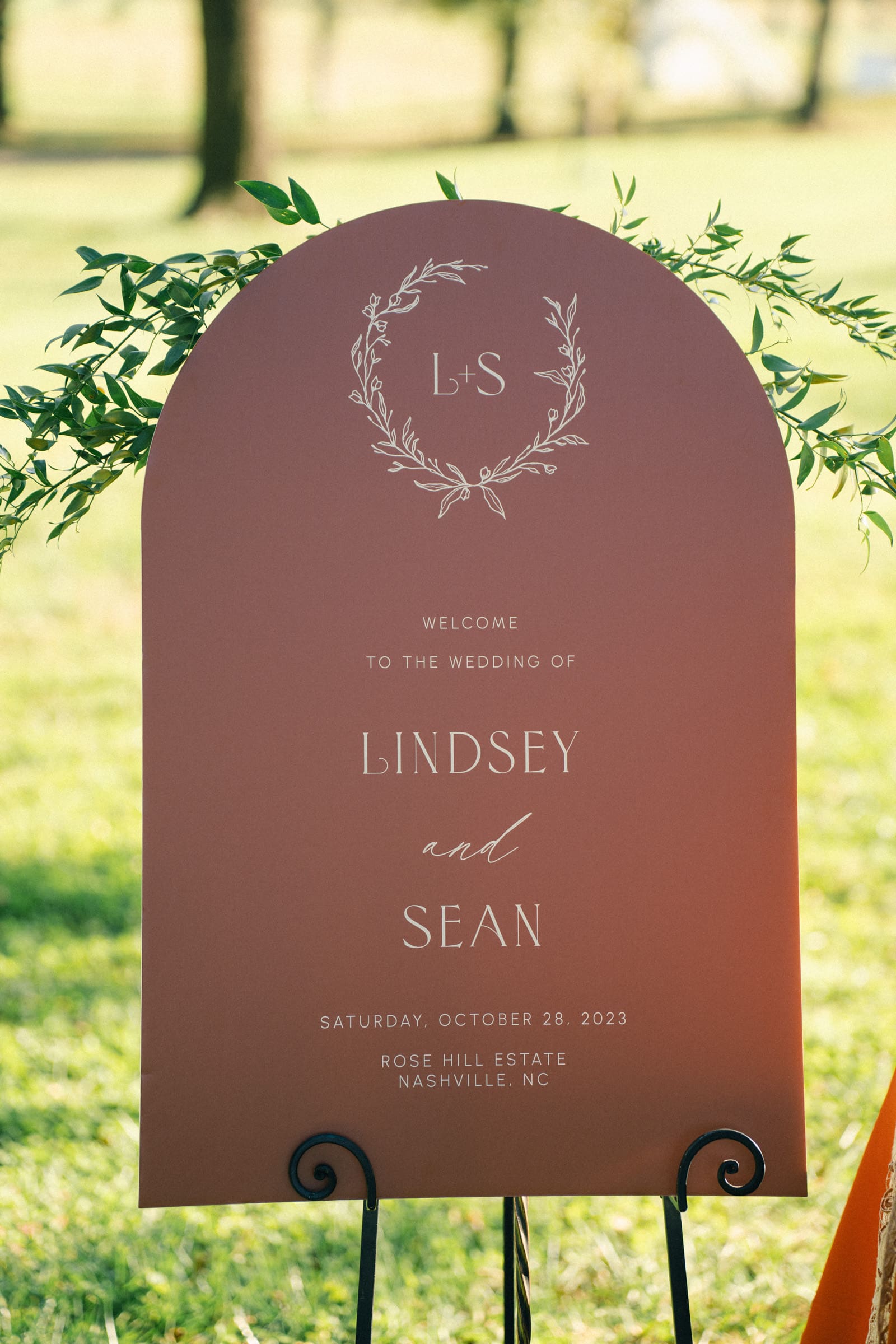 A rust-colored wedding sign with delicate foliage design at the top greets guests: L+S. Welcome to the Rose Hill Estate wedding of Lindsey and Sean. Saturday, October 28, 2023, in Nashville, NC. Mounted on a black stand with greenery in the background.