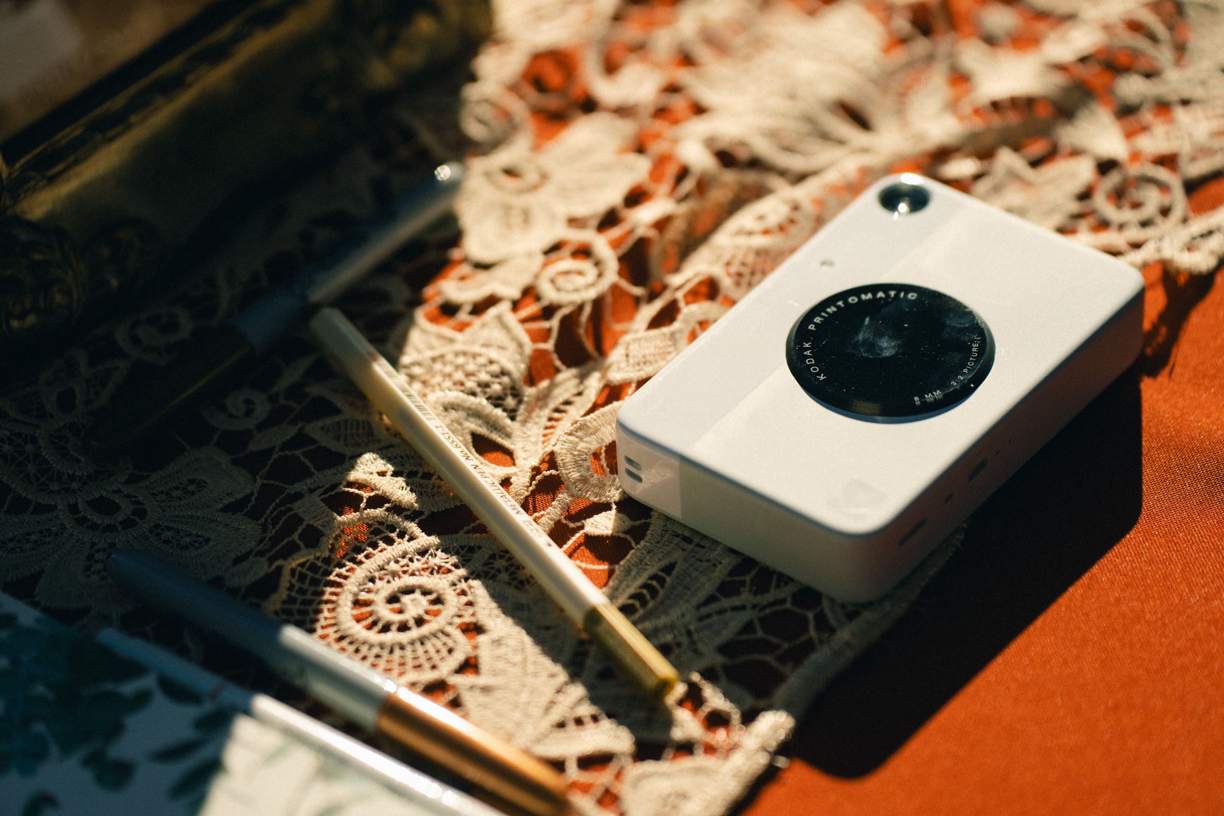 A white instant camera sits on a decorative lace tablecloth, surrounded by pens and a notebook, evoking memories of a Rose Hill Estate wedding. Sunlight casts soft shadows, creating a warm, nostalgic atmosphere.