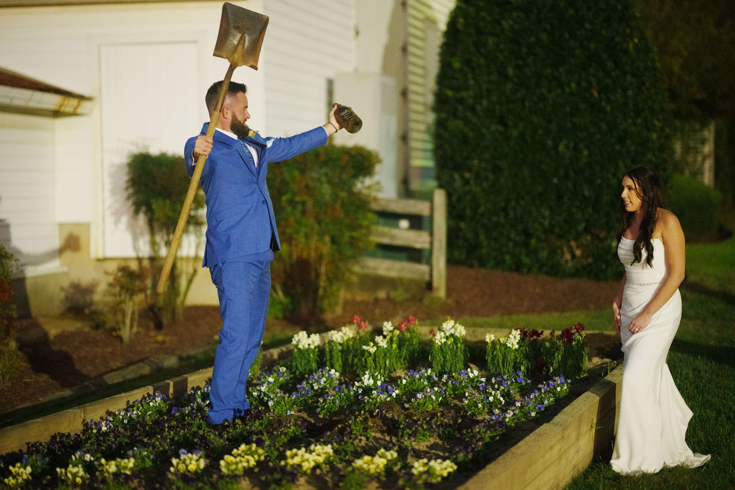 A person in a blue suit stands triumphantly in a garden bed at the Rose Hill Estate, holding a shovel. Another person in a white dress looks on, smiling. The garden is bordered by a wooden frame and filled with colorful flowers, creating the perfect backdrop for this wedding scene.