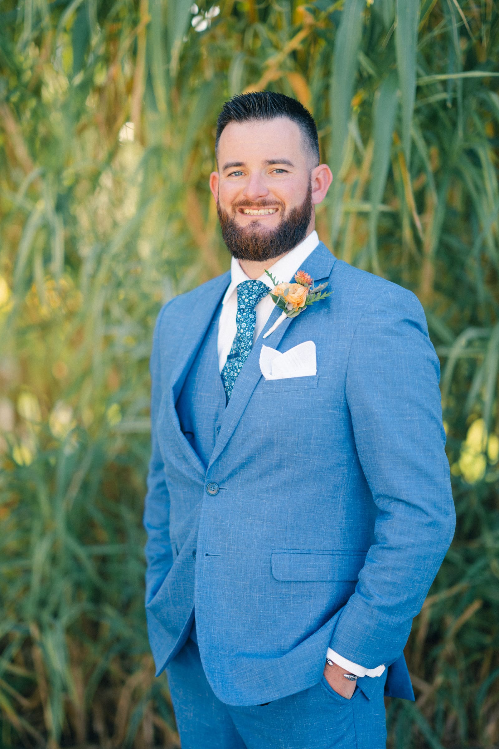 A man in a blue suit with a floral tie stands outdoors at Rose Hill Estate, surrounded by lush green foliage. Sporting a beard and an orange flower boutonniere, he smiles warmly, hands tucked into his pockets.