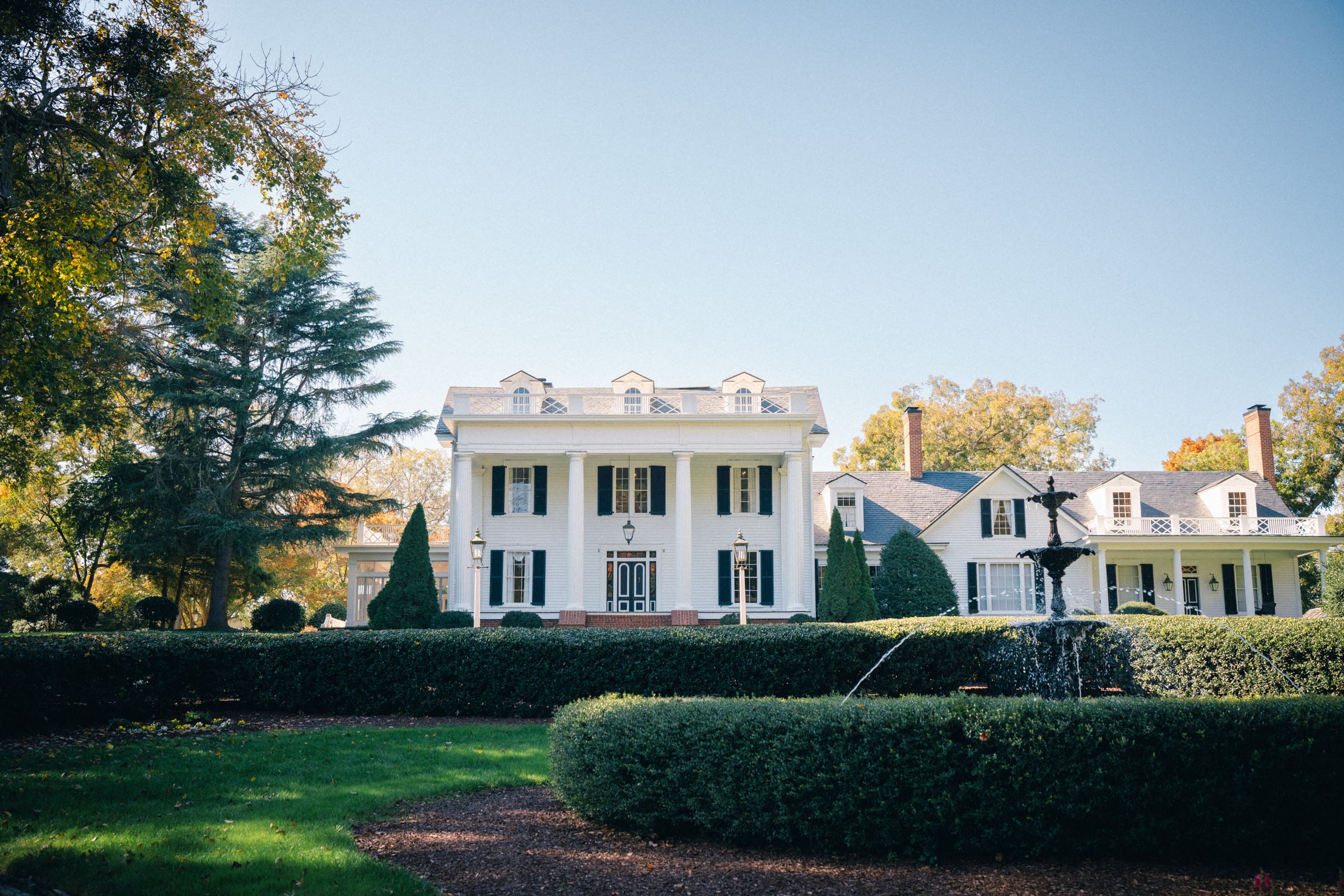 A large, elegant white colonial-style house with columns and black shutters stands amidst neatly trimmed hedges and a manicured lawn, perfect for a Rose Hill Estate wedding. A fountain graces the foreground, and trees frame the scene under a clear blue sky.