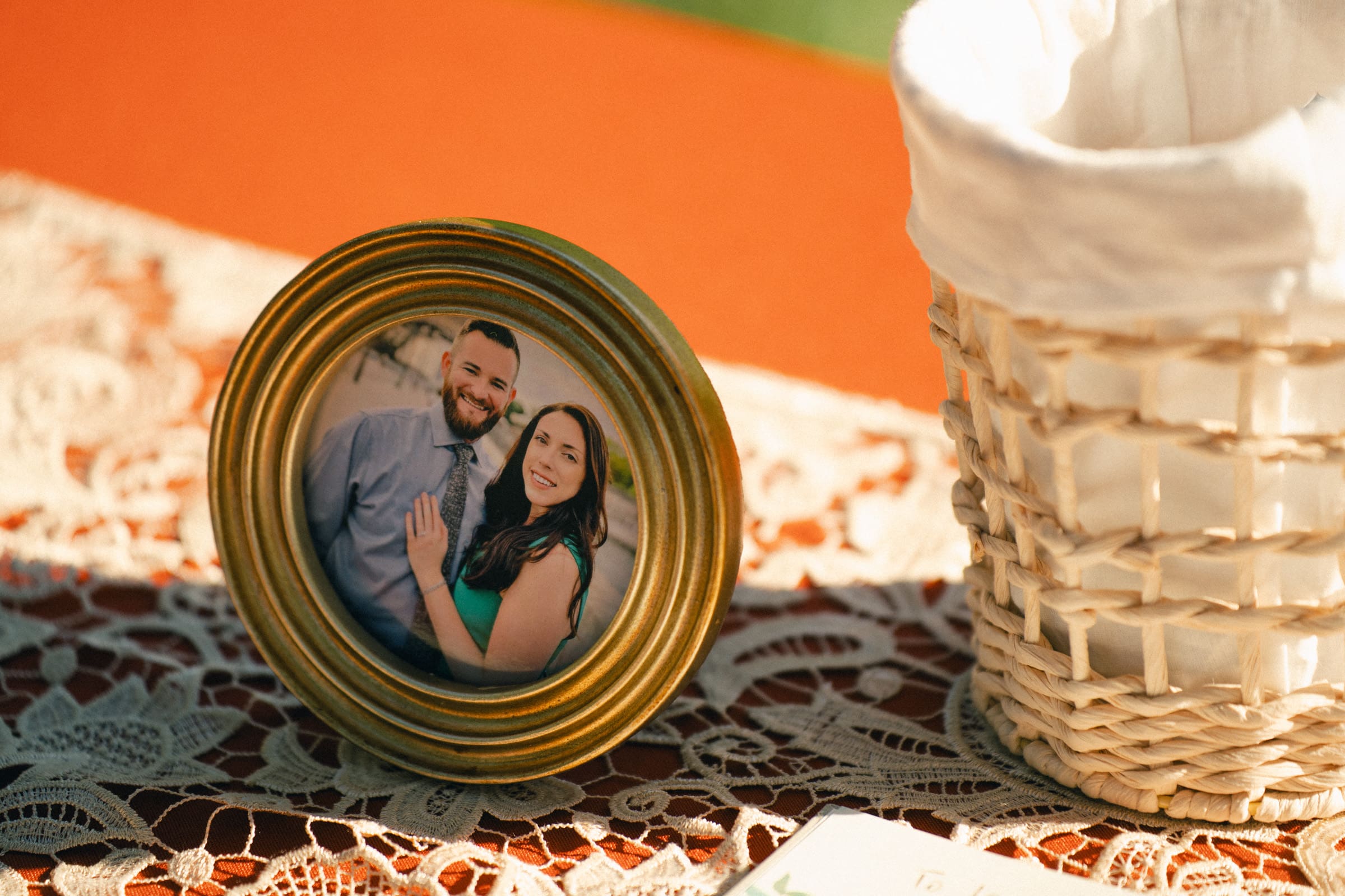 A framed photo of a smiling couple graces the table with a lace tablecloth, evoking memories of a Rose Hill Estate wedding. Beside it sits a woven basket with a white cloth lining, completing the sunny and warm setting.