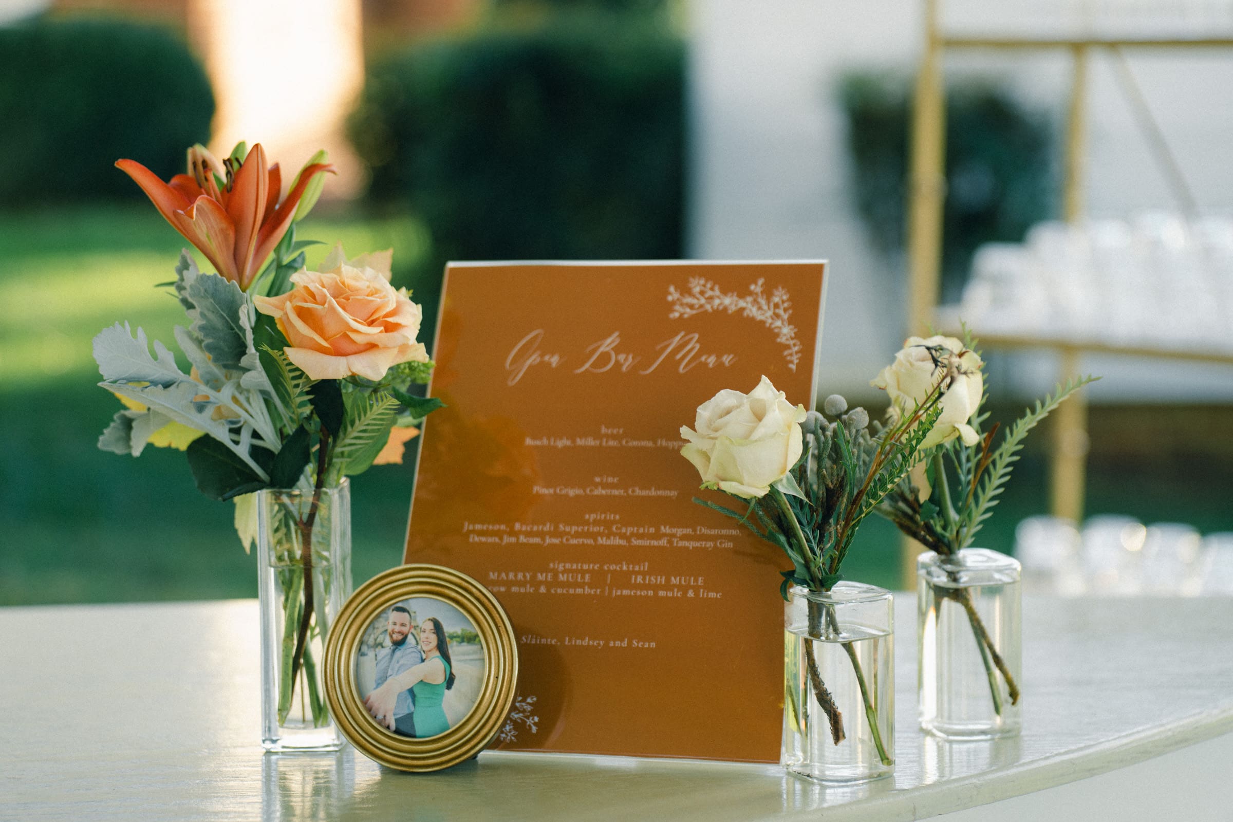 Amidst the lush greenery, a decorative table at the Rose Hill Estate wedding features small floral arrangements and a framed photo. An orange sign with elegant typography lists beverages, including wine, beer, and specialty drinks.