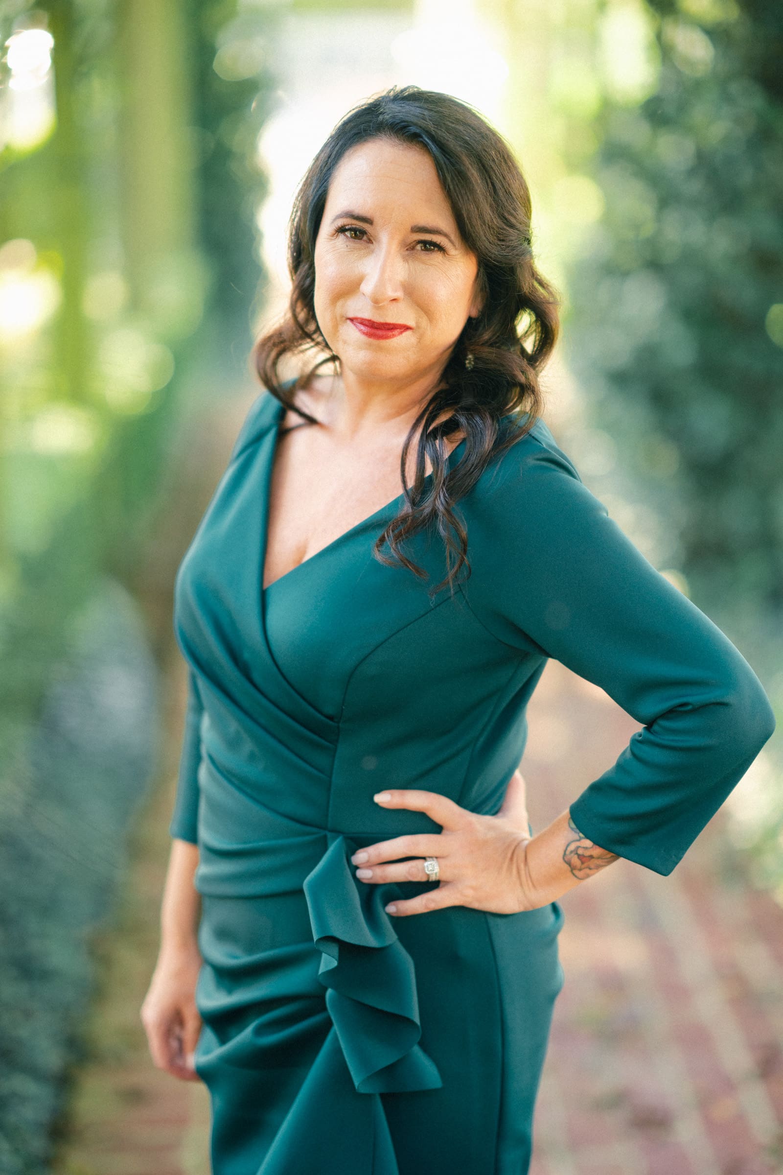 A woman with long, wavy brown hair stands confidently outdoors at Rose Hill Estate. She is wearing a dark green, long-sleeved dress and has a slight smile. The softly blurred greenery in the background evokes the beauty of a serene wedding setting.