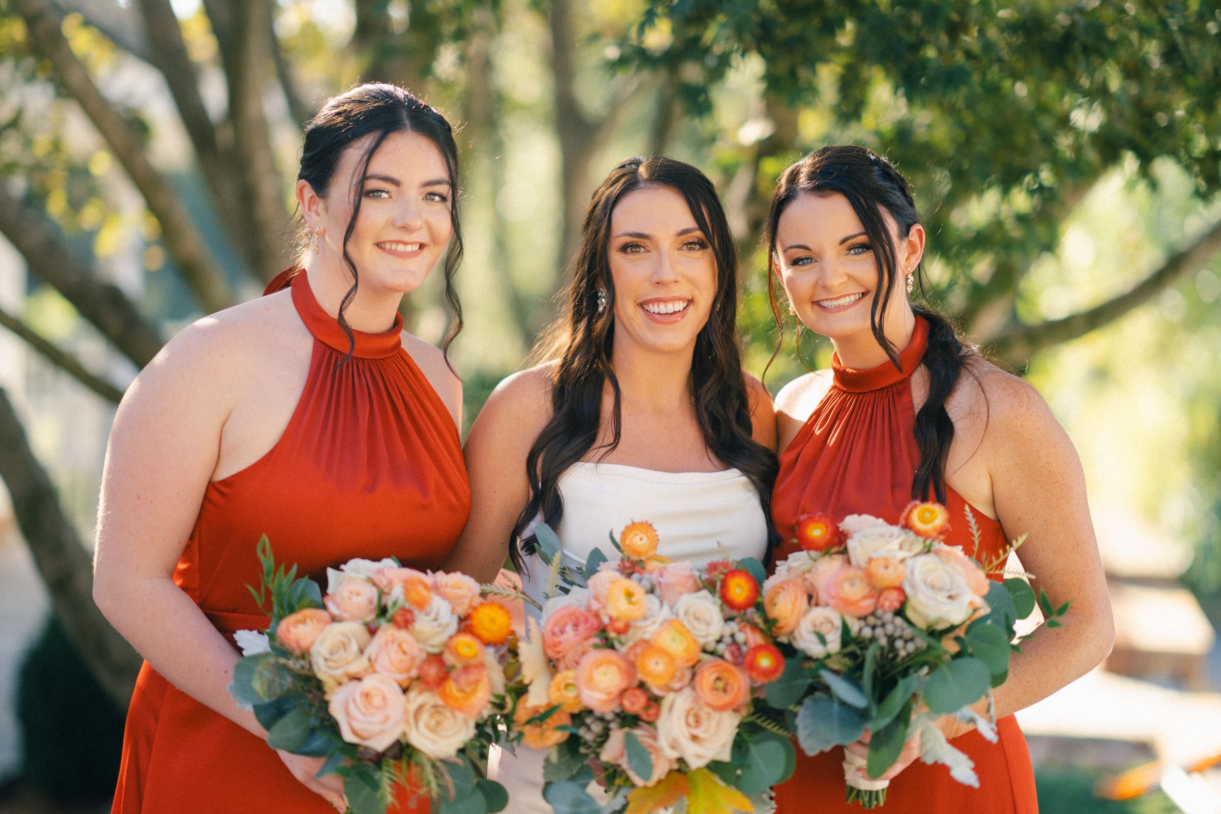 Three women smile in sleeveless red dresses, holding colorful bouquets with roses and greenery. They stand outdoors at Rose Hill Estate, with sunlight filtering through the trees in the background, evoking a dreamy wedding scene.