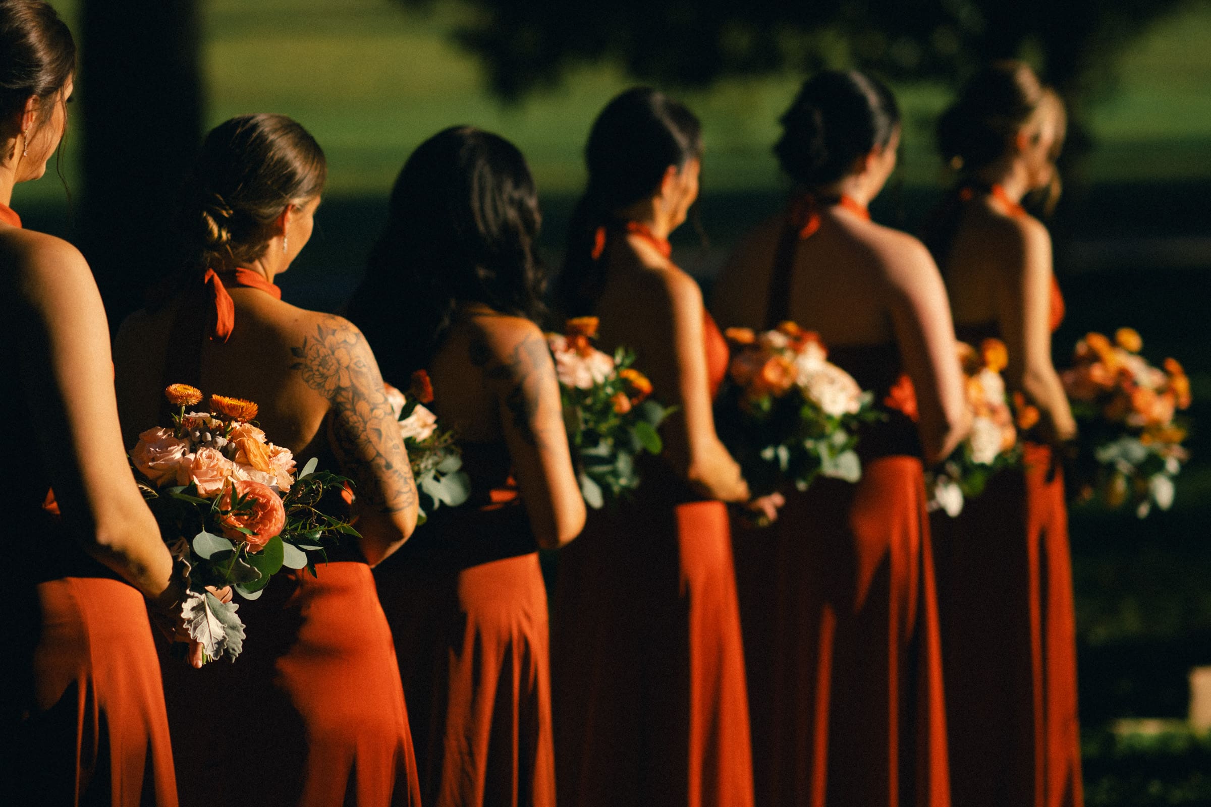 A line of bridesmaids in rust-colored dresses, holding bouquets of orange and white flowers, stand outdoors in the warm sunlight at Rose Hill Estate. The background is blurred greenery, highlighting the elegant and serene atmosphere of this enchanting wedding occasion.