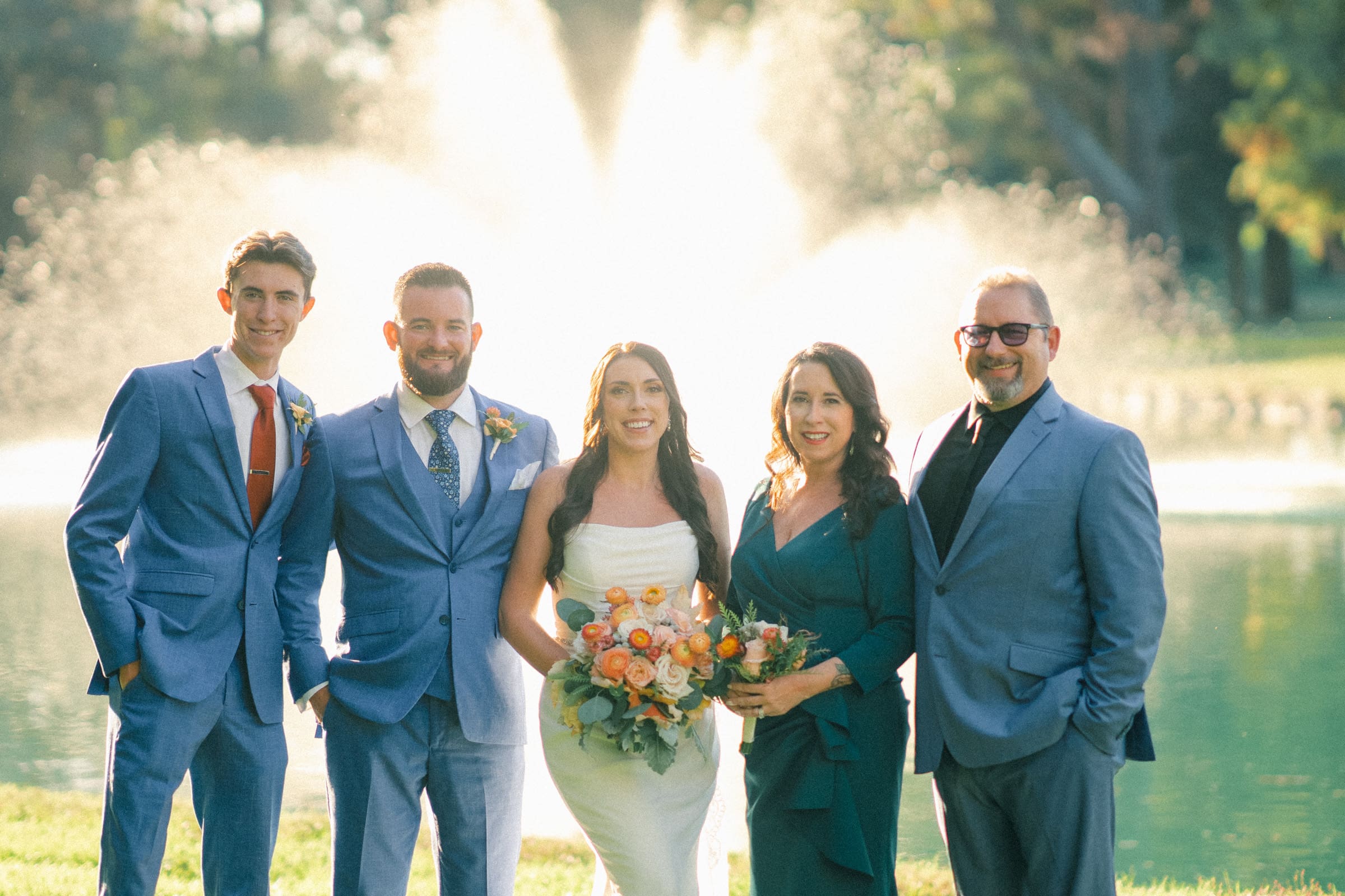 A group of five people, including a bride in a white dress holding a bouquet of flowers, pose in front of a sunlit water fountain at Rose Hill Estate. The others are dressed in blue and green formal attire, smiling warmly at the camera.