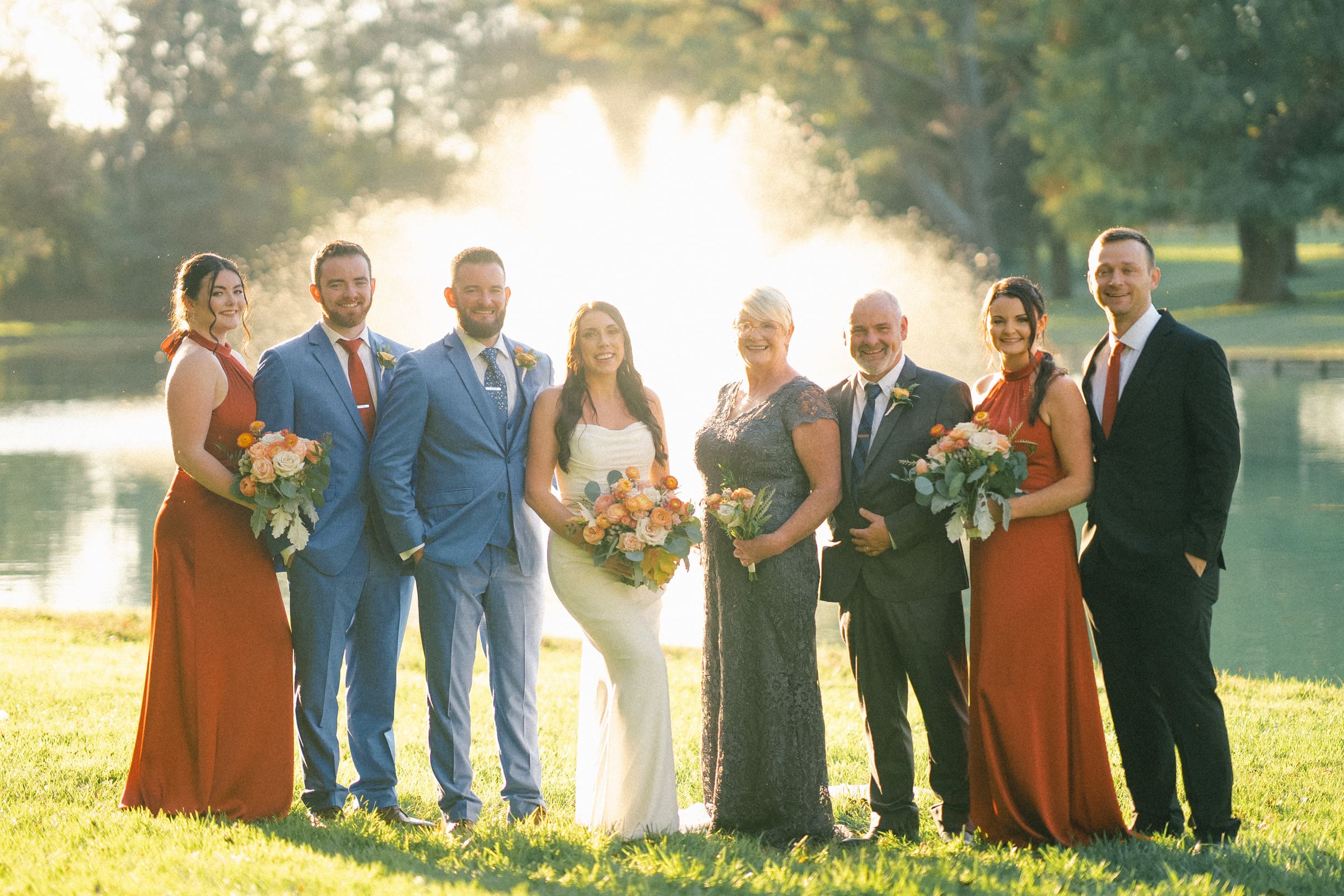 A wedding party at Rose Hill Estate poses in front of a fountain. The group includes four men in suits and four women in dresses, three in burnt orange and one in dark blue. They are all smiling and holding bouquets, with a pond and trees adding to the enchanting backdrop.