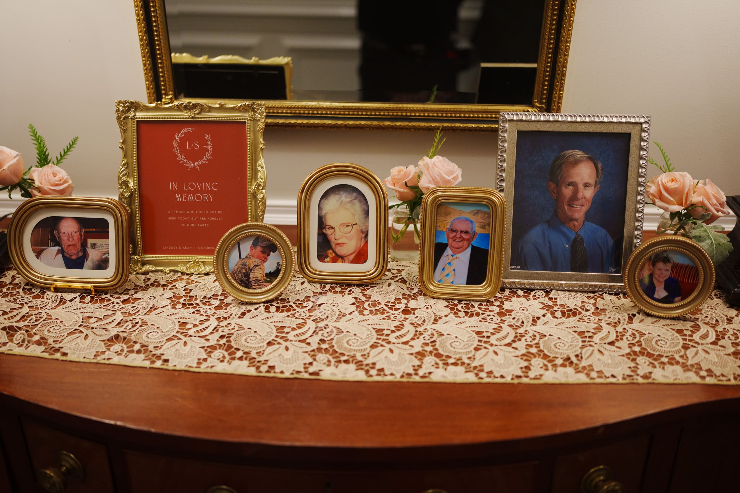 A collection of framed photographs is arranged on a lace-covered wooden dresser, reminiscent of a Rose Hill Estate wedding. The images feature individuals of various ages, with a framed "In Loving Memory" card. Pink roses in vases decorate the scene, while a gold-framed mirror stands gracefully in the background.