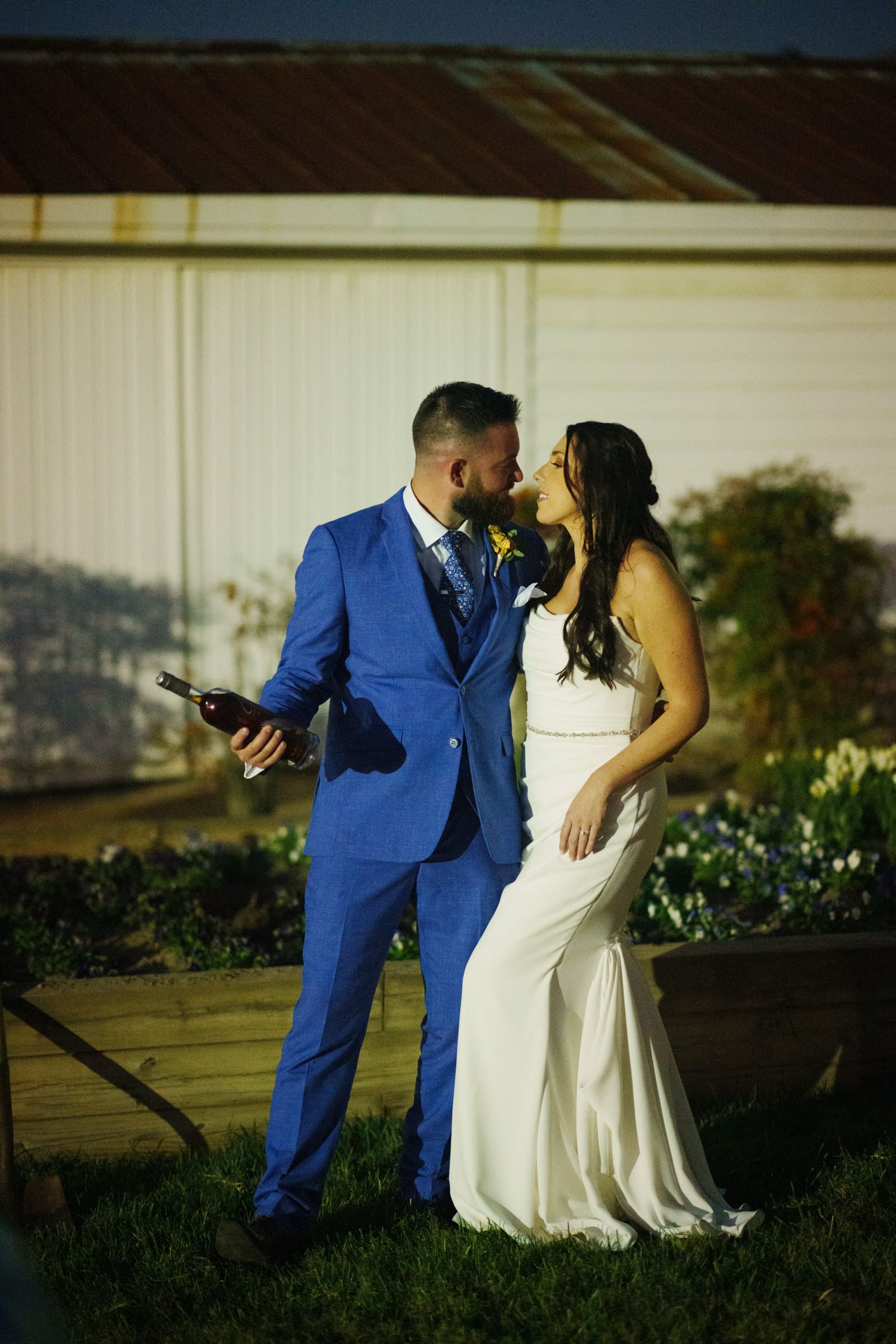 A couple dressed in wedding attire poses outdoors at night at the Rose Hill Estate. The man in a blue suit holds a bottle, while the woman in a white dress smiles at him. They stand on grass with an elegant building and garden in the background, creating a charming scene.
