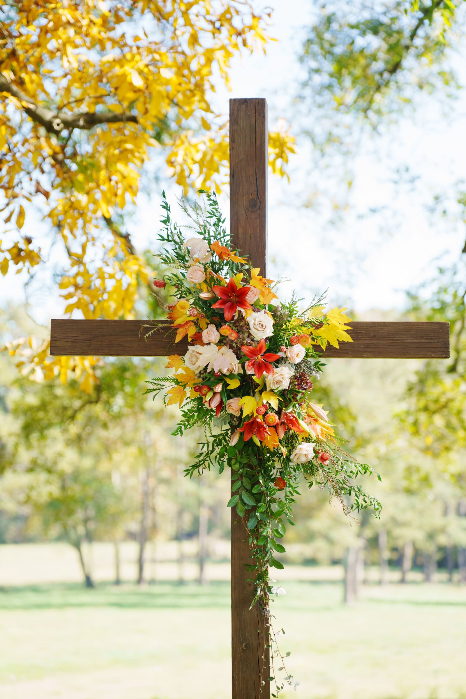 A wooden cross stands outdoors at Rose Hill Estate, adorned with a floral arrangement featuring orange lilies, white and peach roses, and green foliage. The backdrop of a sunny day highlights vibrant autumn trees, setting the perfect scene for a wedding.