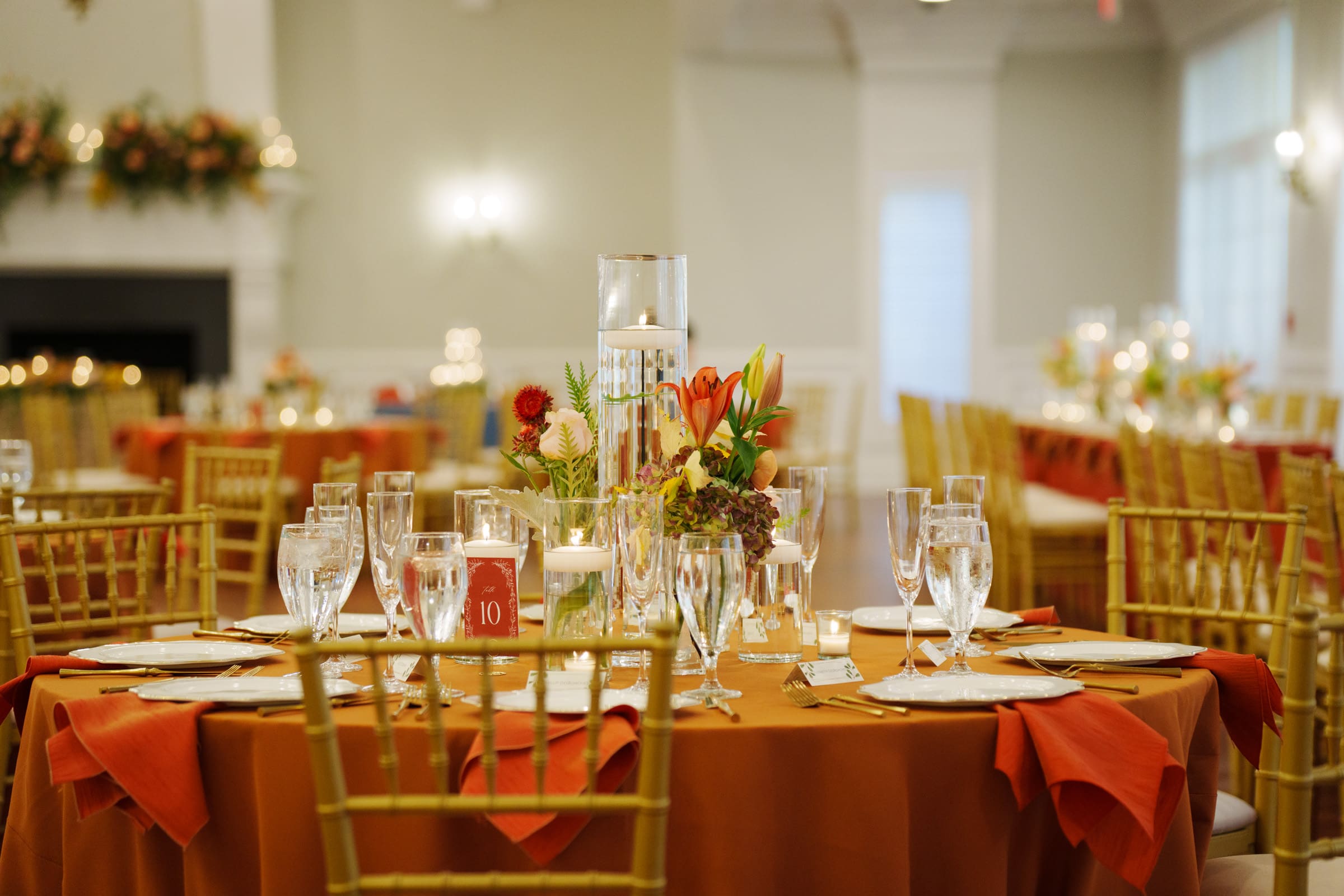 A banquet hall at Rose Hill Estate is set for a wedding event, featuring round tables with orange tablecloths, floral centerpieces, and lit candles. Each table is surrounded by gold chairs, with glassware and plates neatly arranged. The room is softly lit, creating a warm ambiance.