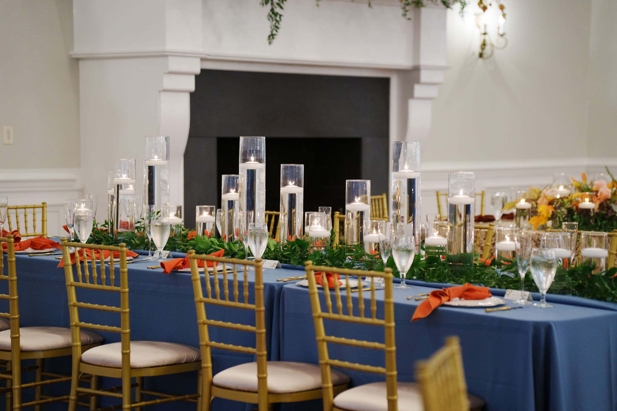 A long table with a blue tablecloth is elegantly set for an event at Rose Hill Estate. Tall glass vases with lit candles, greenery, and orange napkins adorn the table. Gold chairs encircle it, while a fireplace and wall sconces complete the enchanting backdrop.
