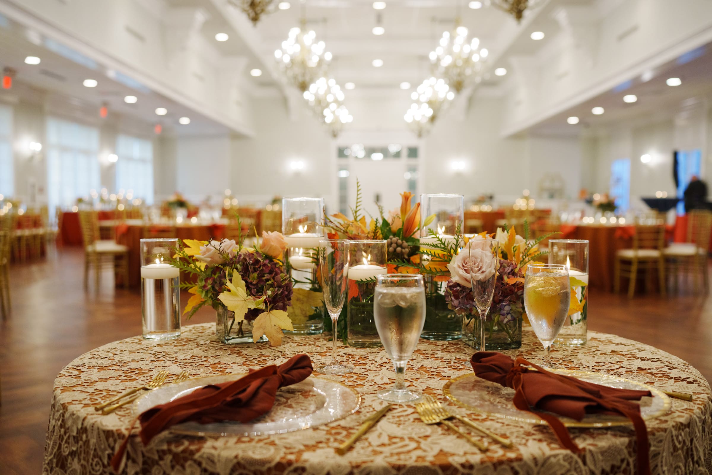 An elegantly set dining table with floral centerpieces, candles, and glassware is in focus. The background shows a large, brightly lit banquet hall with chandeliers at Rose Hill Estate, perfectly set for a formal wedding event.