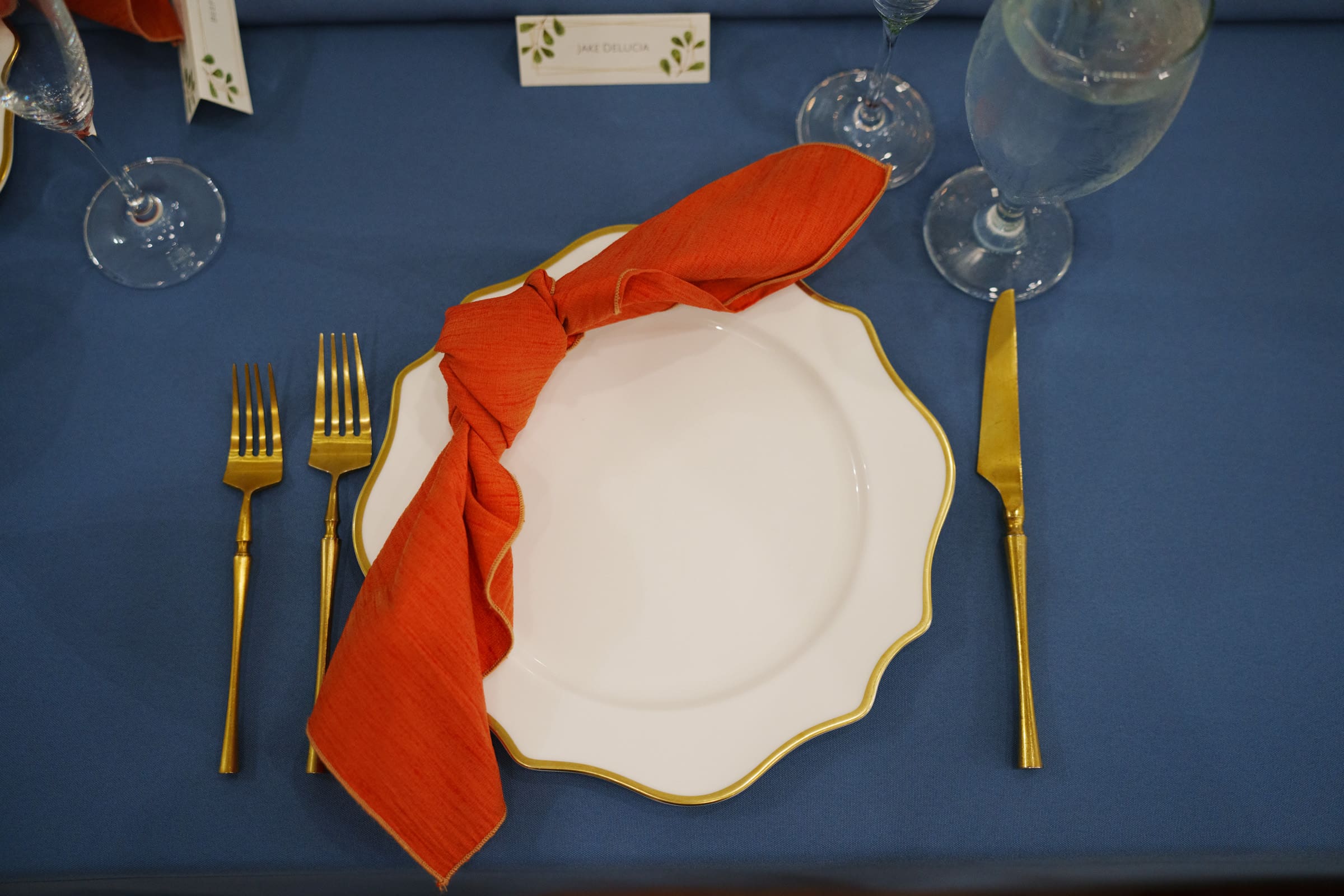 A place setting for a Rose Hill Estate wedding showcases a white scalloped plate with gold edges, an orange napkin artistically wrapped around, and gold-colored cutlery. A name card rests on the blue tablecloth alongside a glass of water, epitomizing elegance and charm.