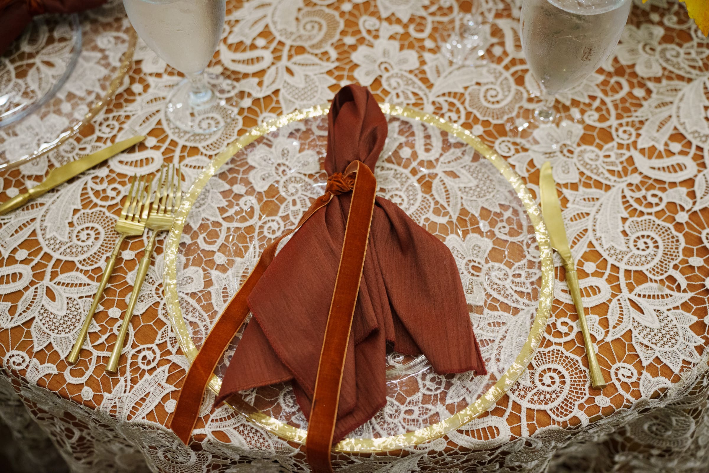 An elegantly set table at a Rose Hill Estate wedding boasts a lace tablecloth featuring a clear plate with a rust-colored napkin tied with a ribbon, surrounded by gold utensils and wine glasses filled with water.