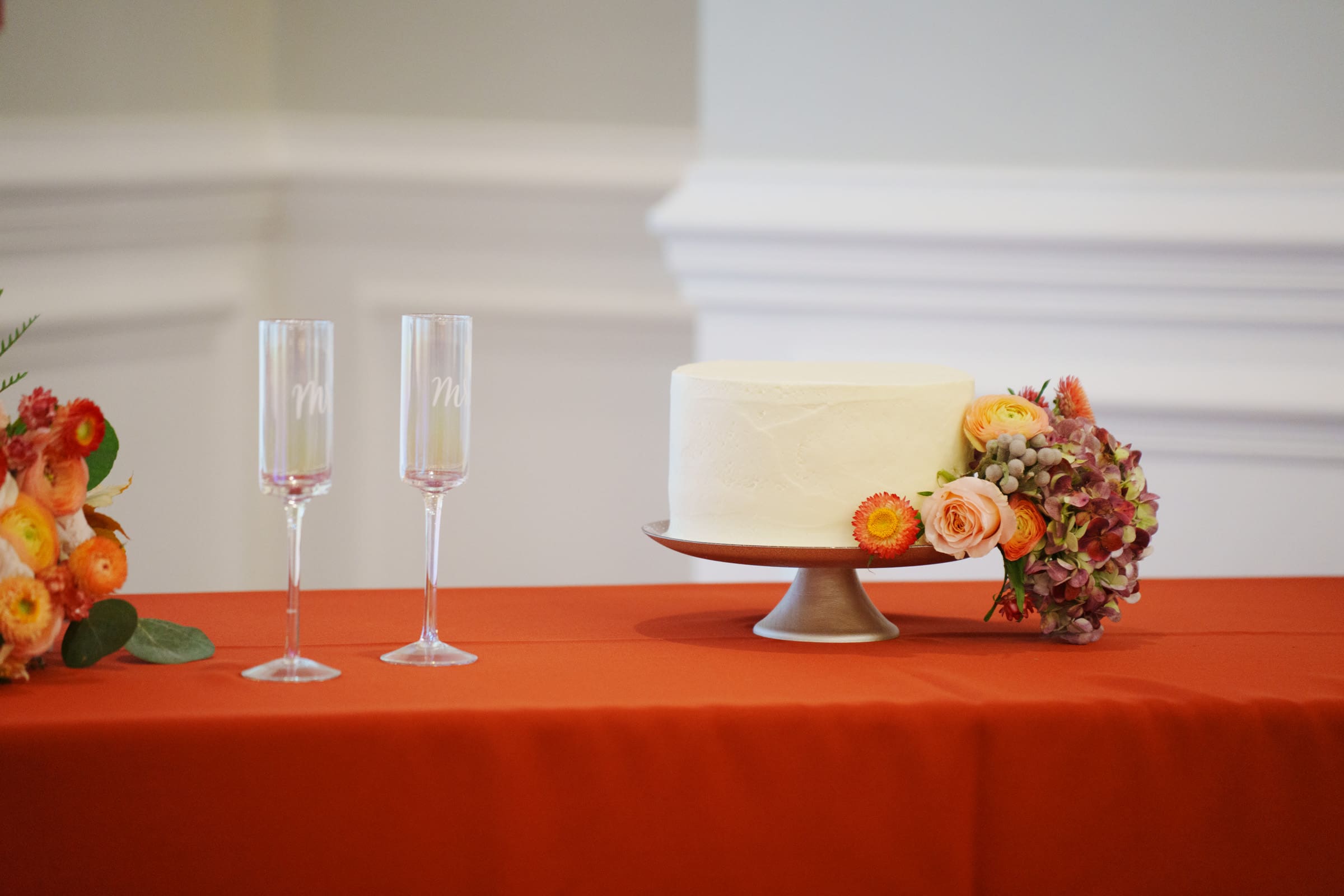 A round white cake on a stand sits on an orange tablecloth, surrounded by colorful flowers and two champagne flutes with initials at a Rose Hill Estate wedding. The elegant backdrop features a white wall with molding, enhancing the sophisticated setting.