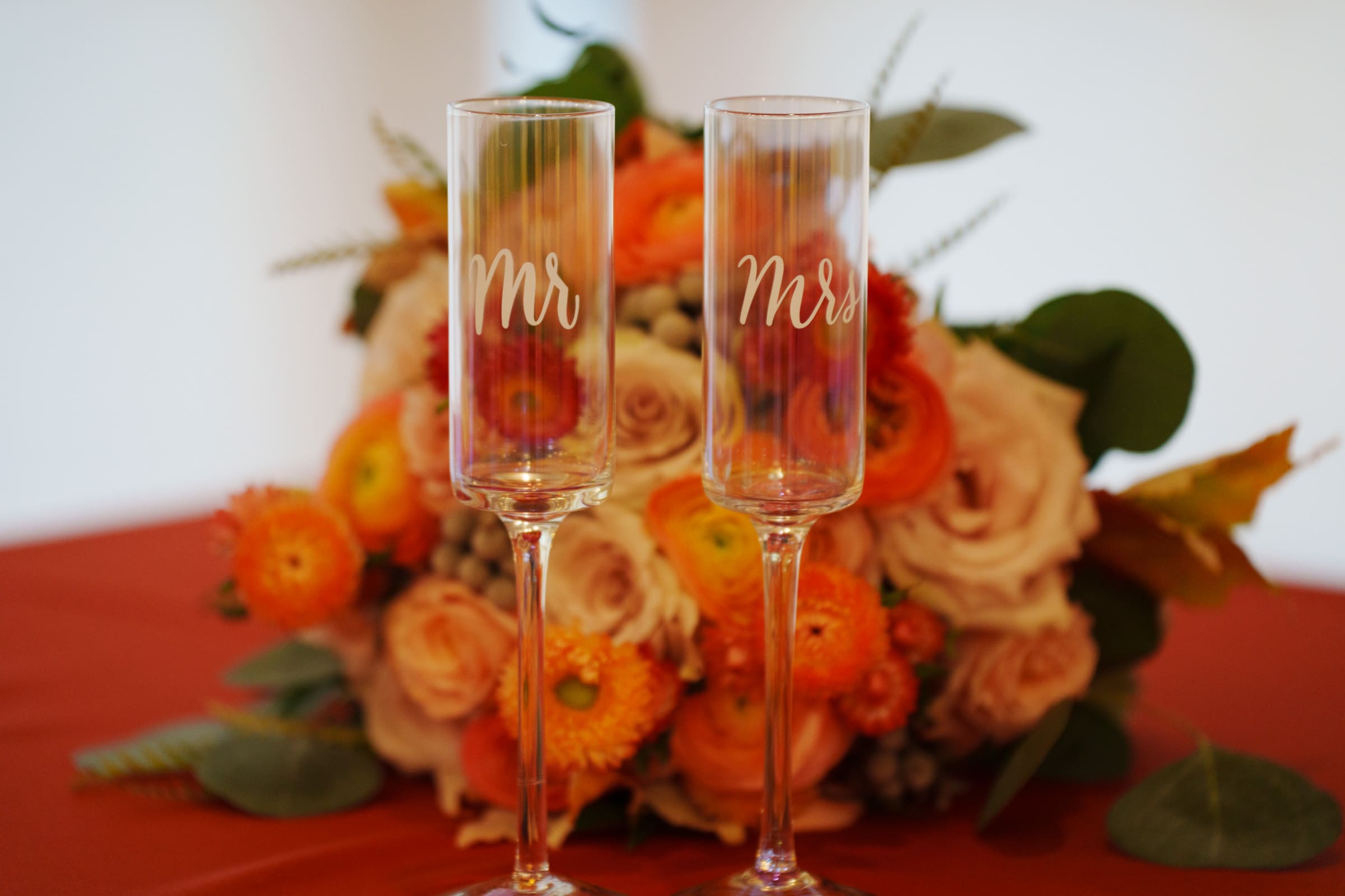 Two champagne flutes with Mr. and Mrs. etched on them are elegantly arranged in front of a bouquet of orange and pink flowers with green leaves, set on a red tablecloth, embodying the romantic charm of a Rose Hill Estate wedding.