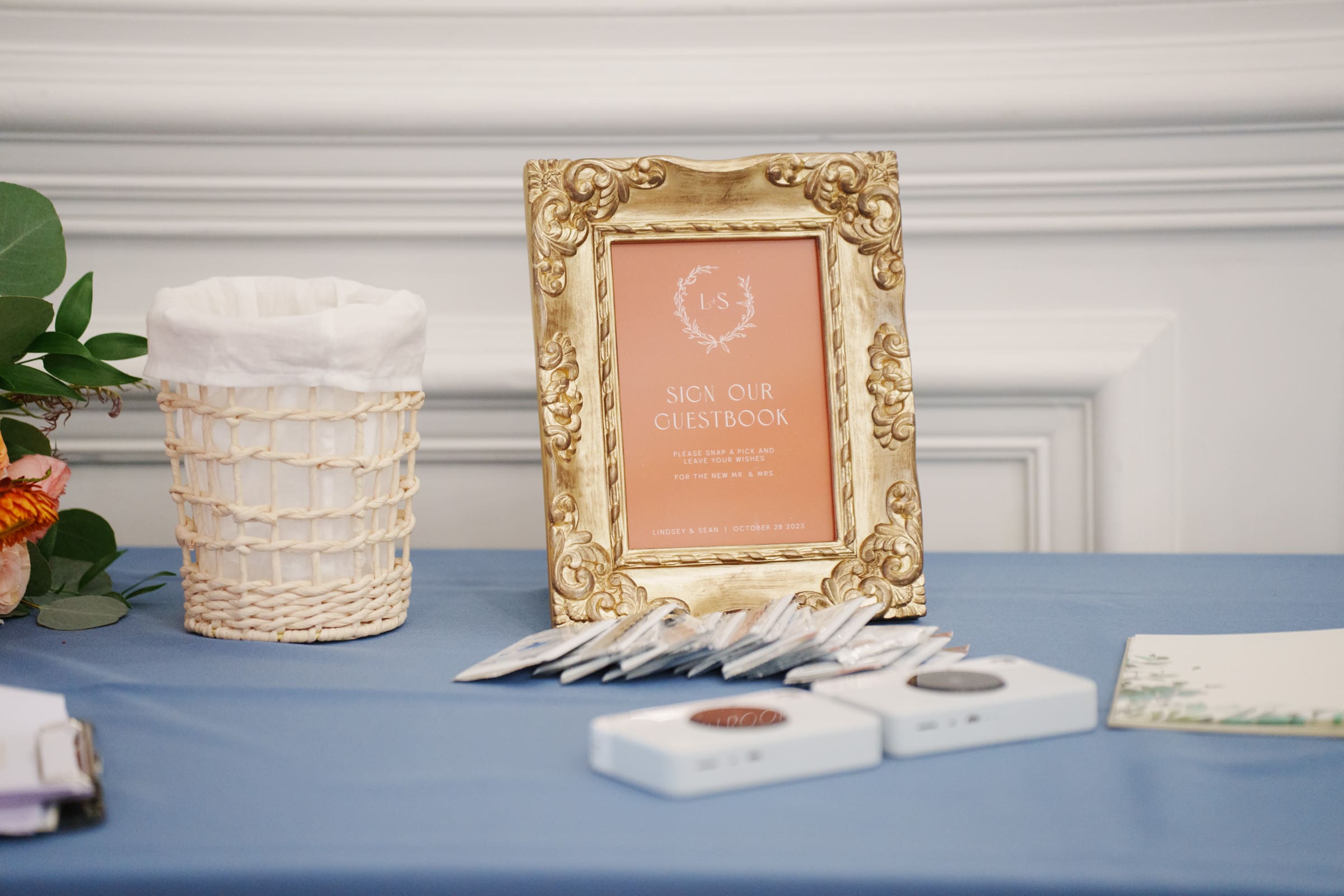 A table at the Rose Hill Estate wedding, draped in a blue cloth, showcases a gold frame with a 'Sign Our Guestbook' sign adorned with elegant details. Beside it, find a woven basket and charming arrangements of small packets and card boxes with flowers peeking through.