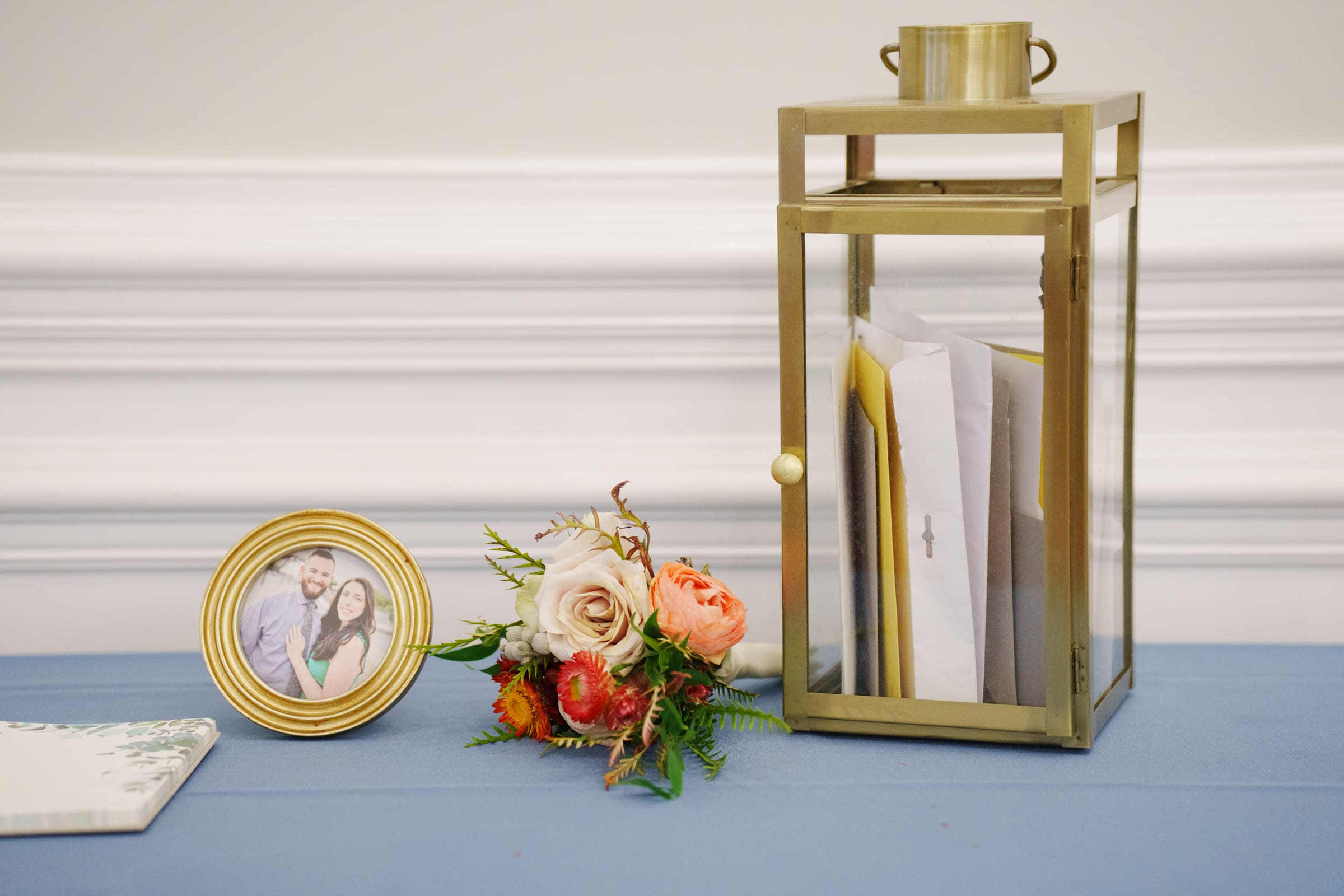 A gold-framed photo of a smiling couple graces the table at the Rose Hill Estate wedding, alongside a small bouquet and a gold lantern cradling envelopes. The setting is completed by a light blue tablecloth against the elegant backdrop of a white wainscoted wall.