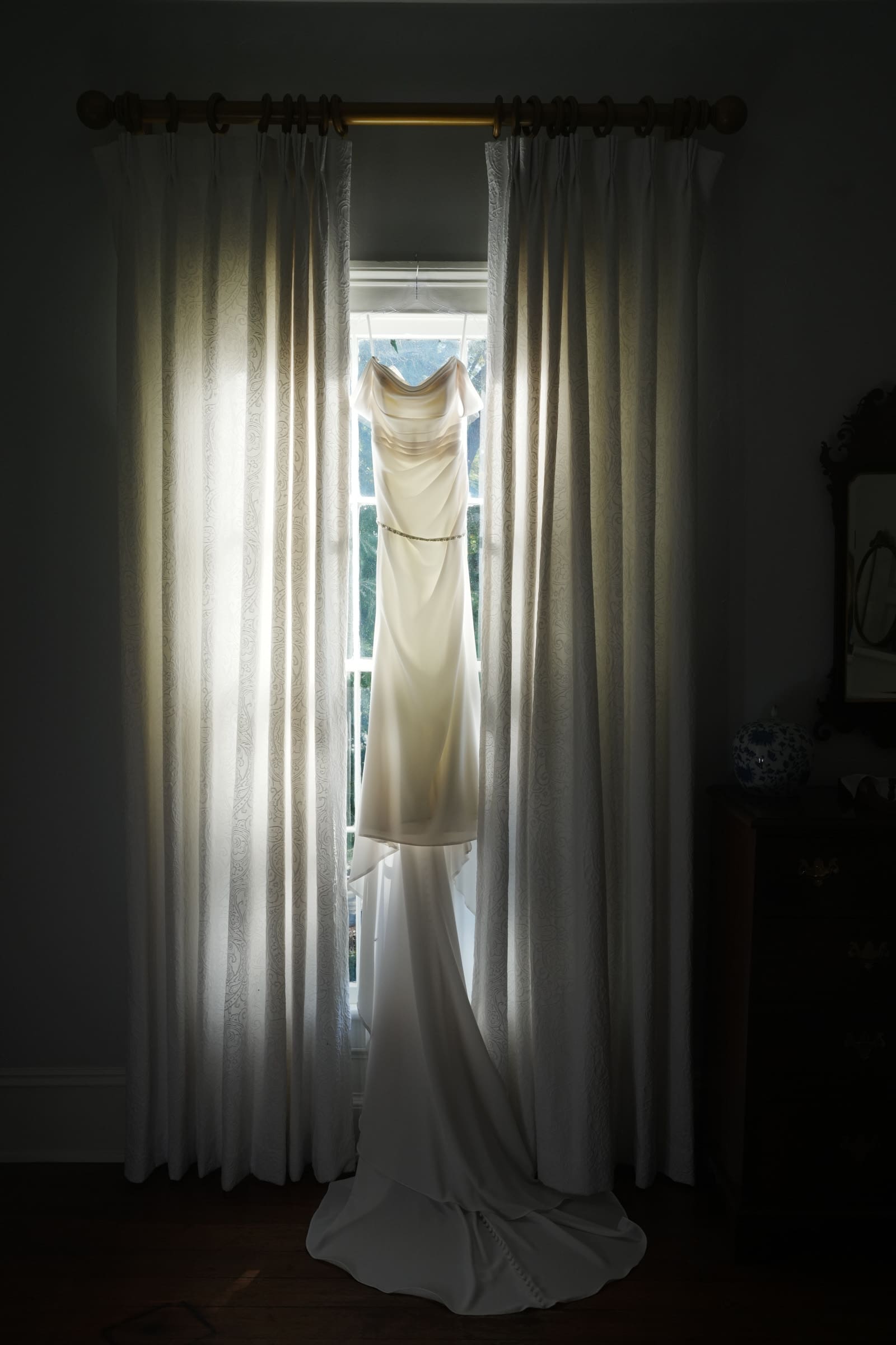 A white wedding dress with a long train elegantly hangs in front of a window, framed by softly lit curtains at Rose Hill Estate. The sunlight gently illuminates the fabric, creating a serene and romantic ambiance in the room.