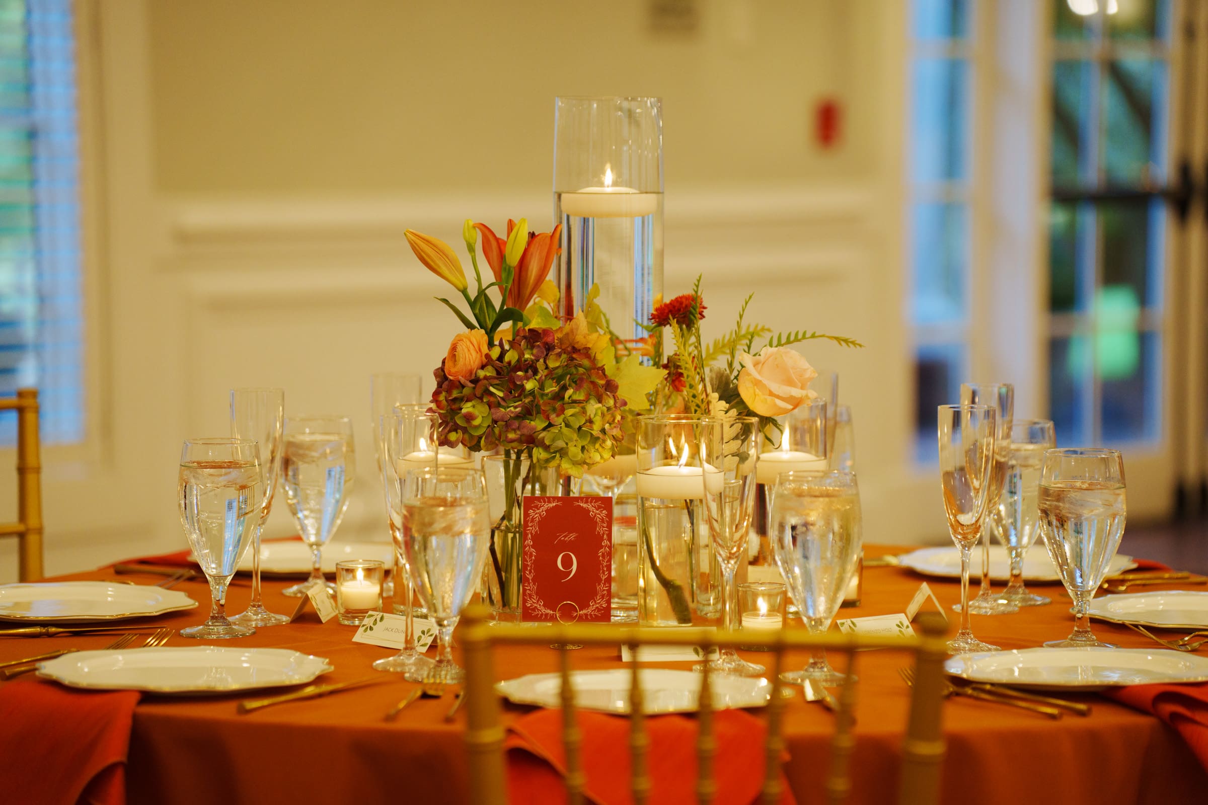 A beautifully set dining table at the charming Rose Hill Estate wedding features a floral centerpiece, candles, and settings for eight. The orange tablecloth perfectly complements the atmosphere, with a red table number card marked 9. Glassware and plates are arranged neatly around the centerpiece.