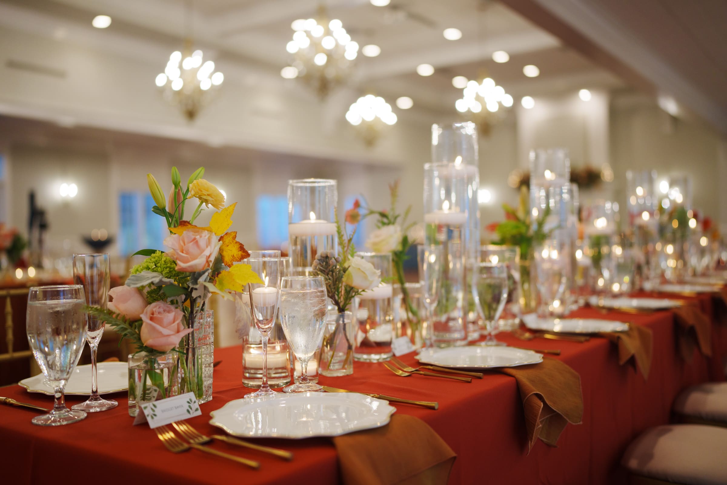 A beautifully set dining table at the Rose Hill Estate wedding features red tablecloths and elegant place settings with white plates, gold utensils, and floral centerpieces. Candles in glass holders add a warm glow, while chandeliers illuminate the softly lit room for a magical ambiance.