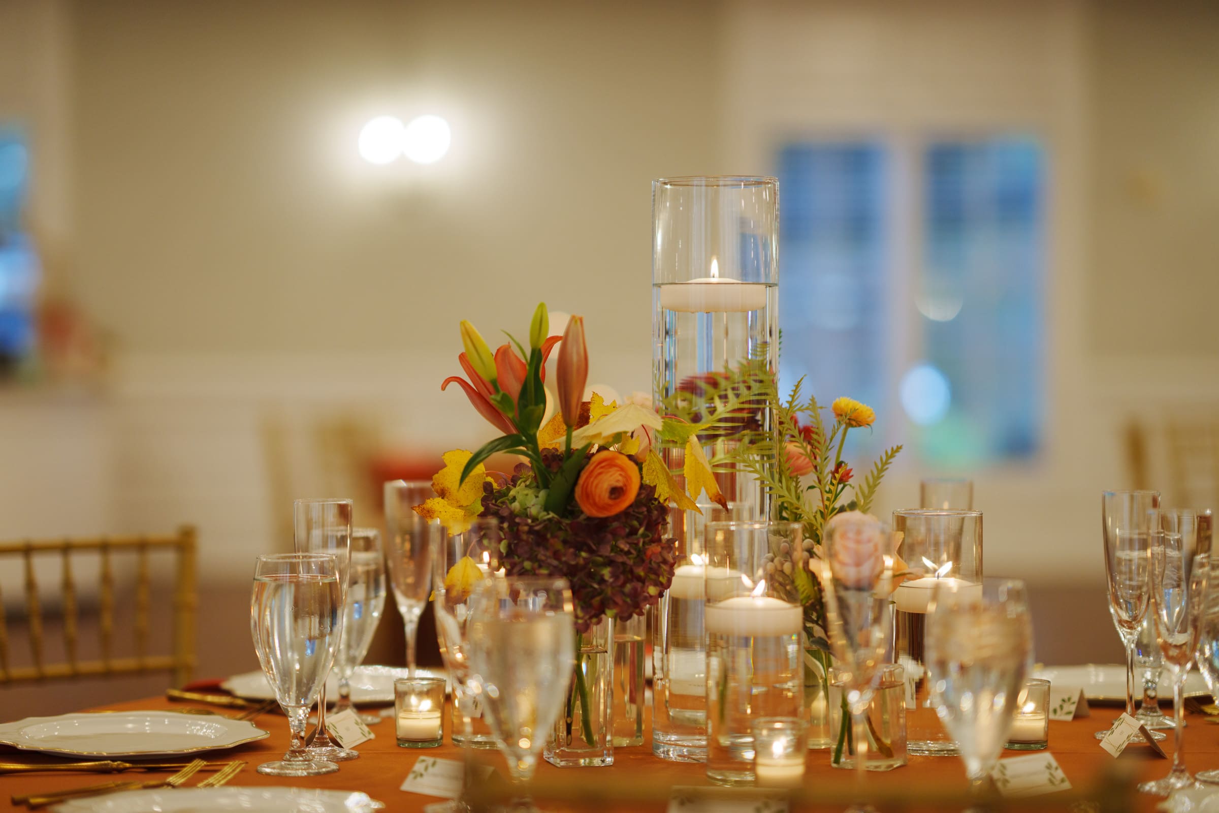 A table elegantly set for a formal event at Rose Hill Estate, featuring a centerpiece with colorful flowers, floating candles, and clear glassware. The background is softly blurred, creating a warm and inviting atmosphere perfect for a wedding reception.