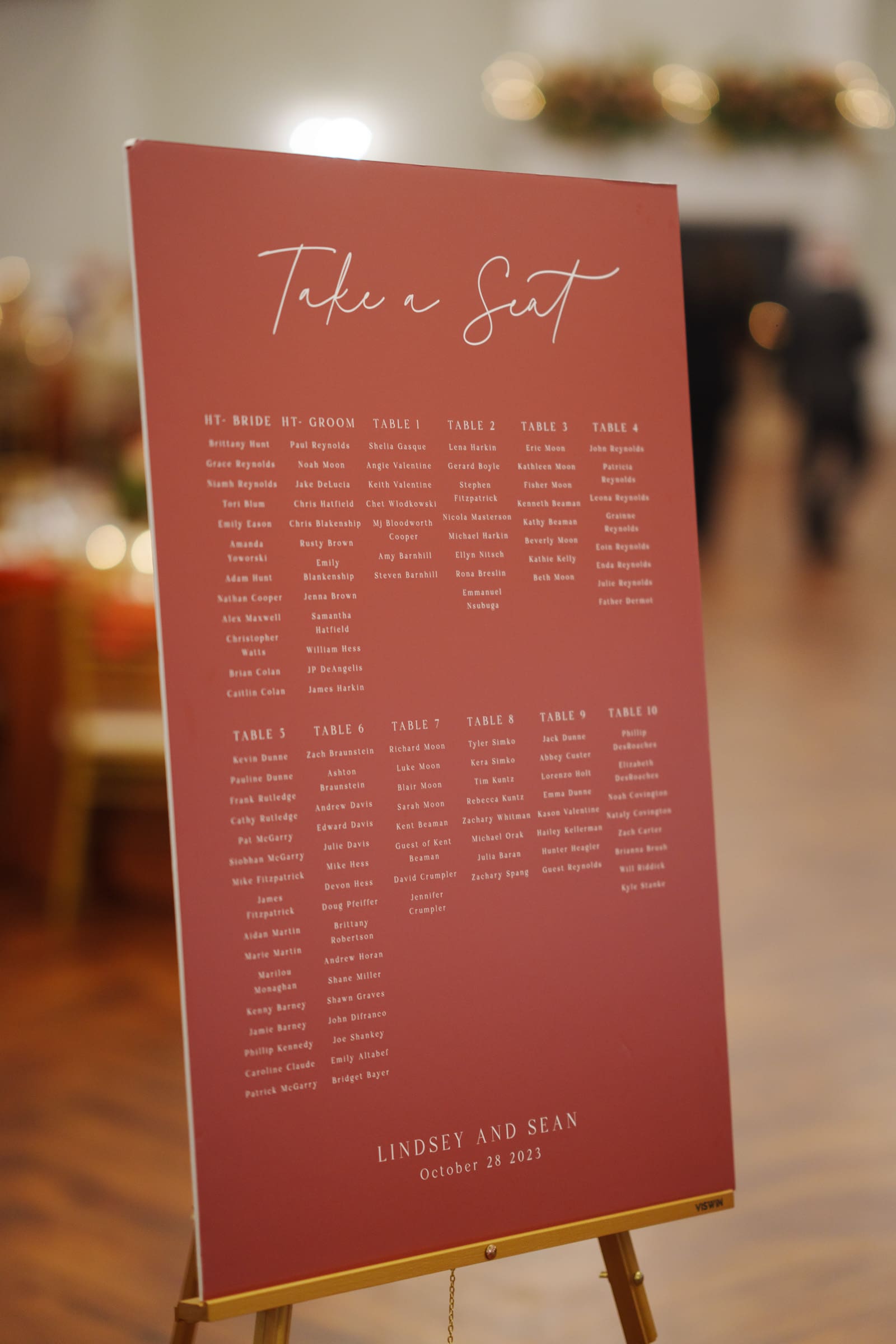 A red wedding seating chart poster titled Take a Seat lists guest names under tables 1-10. It's placed on a stand in the warmly lit reception hall of Rose Hill Estate. The bottom of the poster reads Lindsey and Sean, October 28, 2023.