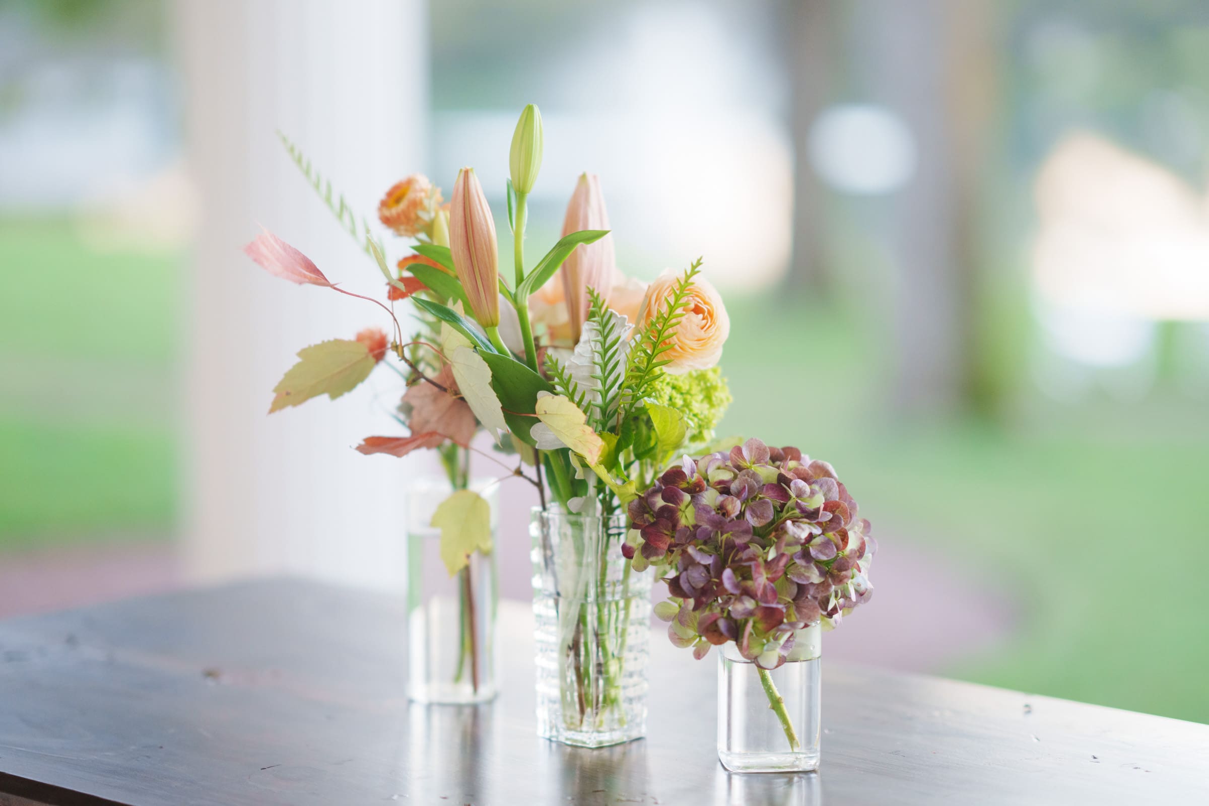 Three vases with floral arrangements sit elegantly on a wooden table, perfect for a Rose Hill Estate wedding. The first vase contains pink and orange blooms, the second has a mix of green and peach flowers, and the third holds a cluster of purple hydrangeas. A blurred outdoor background sets the scene.