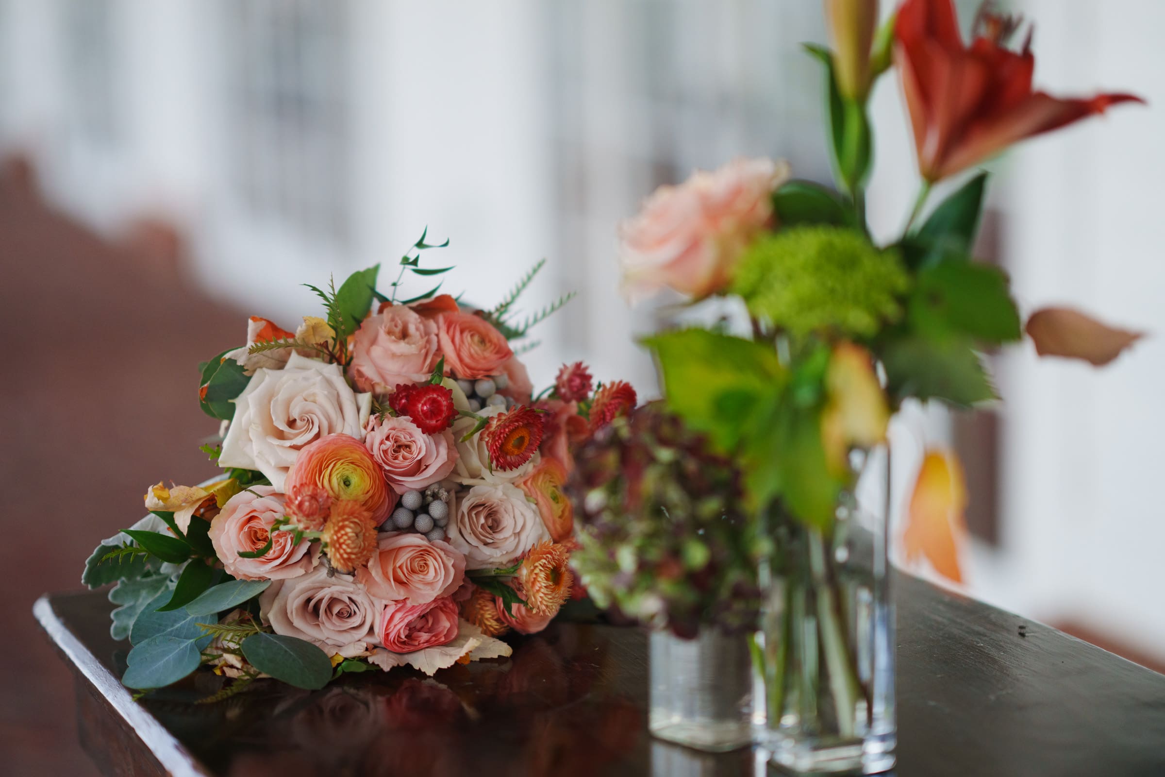 A bouquet of roses and assorted flowers in shades of pink, orange, and white rests on a wooden surface. Next to it, two glass vases hold additional flowers with green and orange blooms. The scene evokes the elegance of a Rose Hill Estate wedding, with its softly blurred background enhancing the charm.