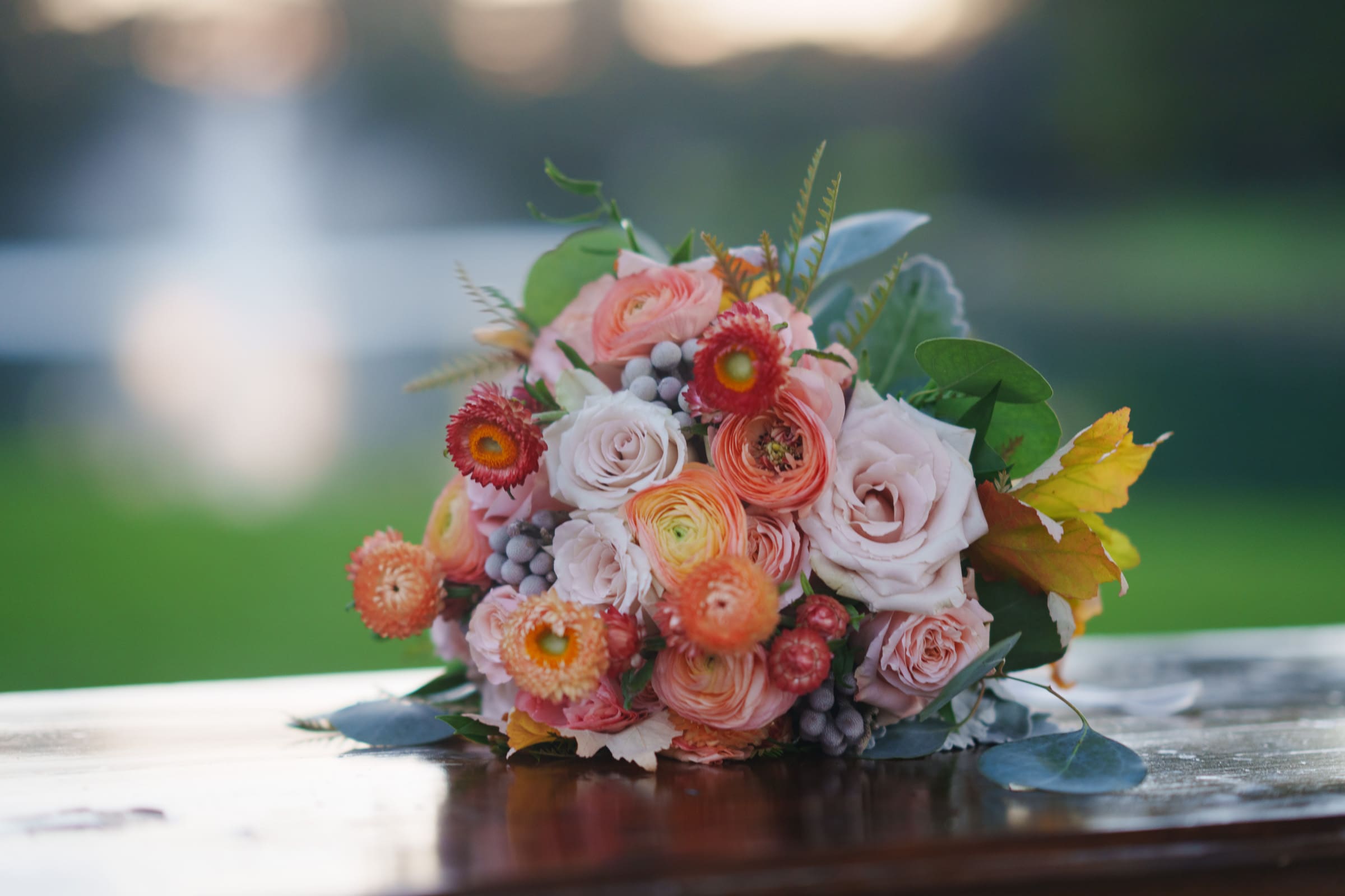 A vibrant bouquet of roses and assorted flowers with pink and orange tones rests on a wooden surface, capturing the elegance of a Rose Hill Estate wedding. Green leaves and a blurred natural background create a serene setting.