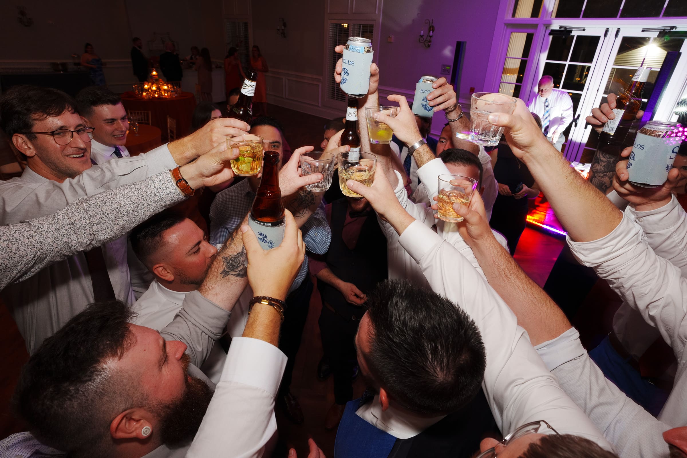 A group of people raise various drinks in a celebratory toast at a Rose Hill Estate wedding. Dressed in formal attire, they enjoy the warm and festive indoor ambience, with colorful lights creating a joyous backdrop to this elegant celebration.