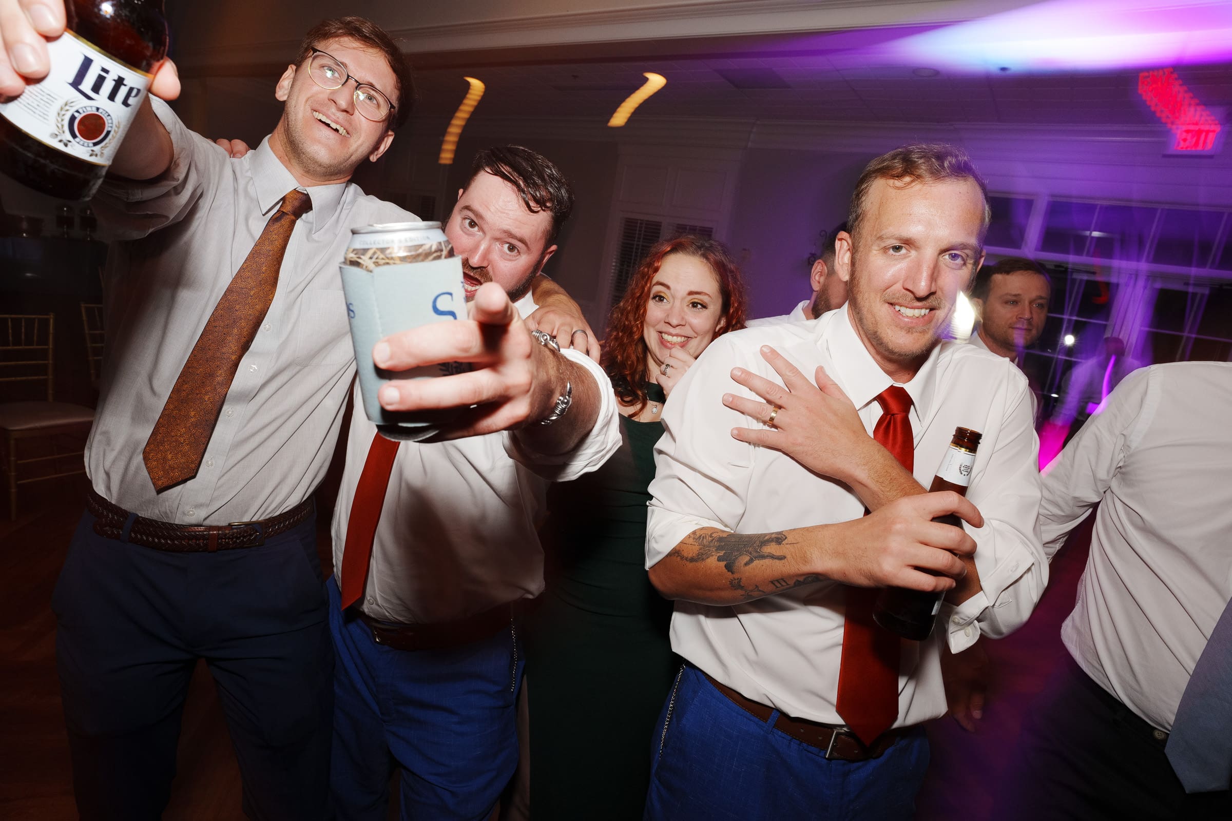 A group of people in formal attire are celebrating on the dance floor at a Rose Hill Estate wedding, holding drinks and smiling. Colorful lights create a festive atmosphere.