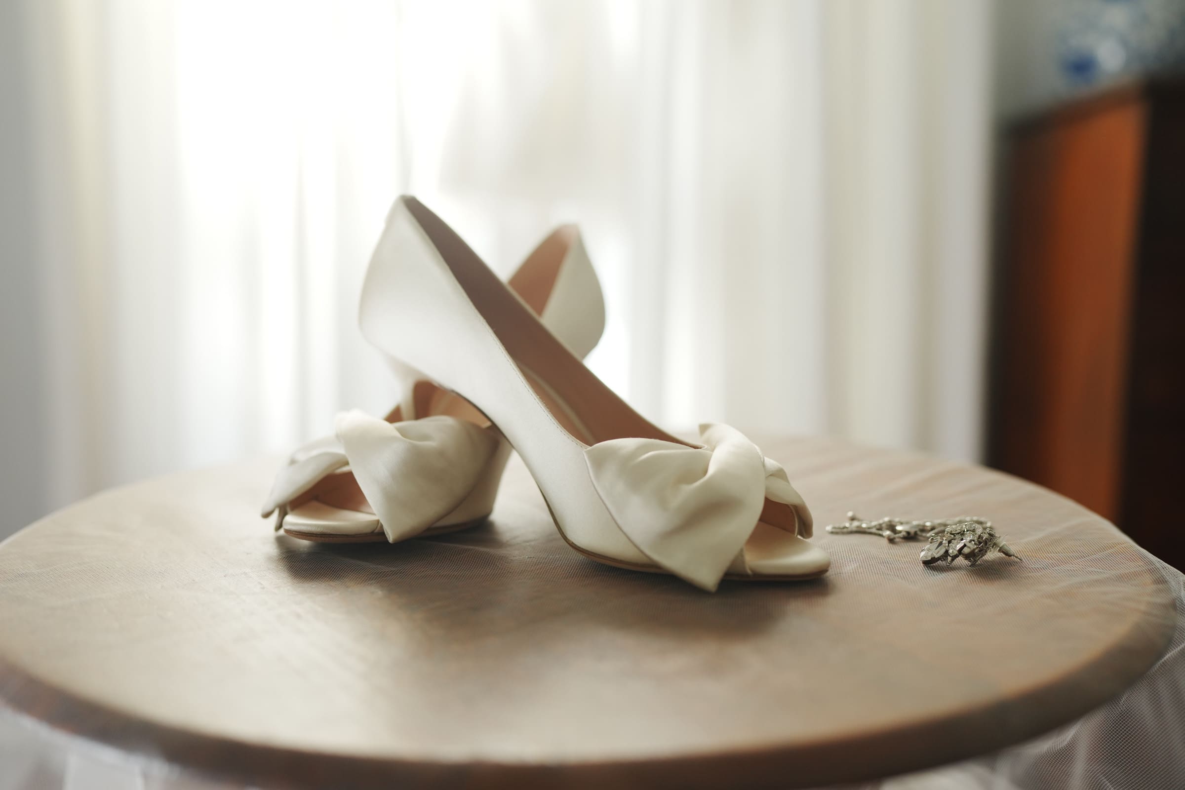 A pair of elegant white satin peep-toe shoes with bow accents are displayed on a wooden surface at the Rose Hill Estate wedding. The background features softly blurred, sheer white curtains, creating a gentle and dreamy atmosphere.