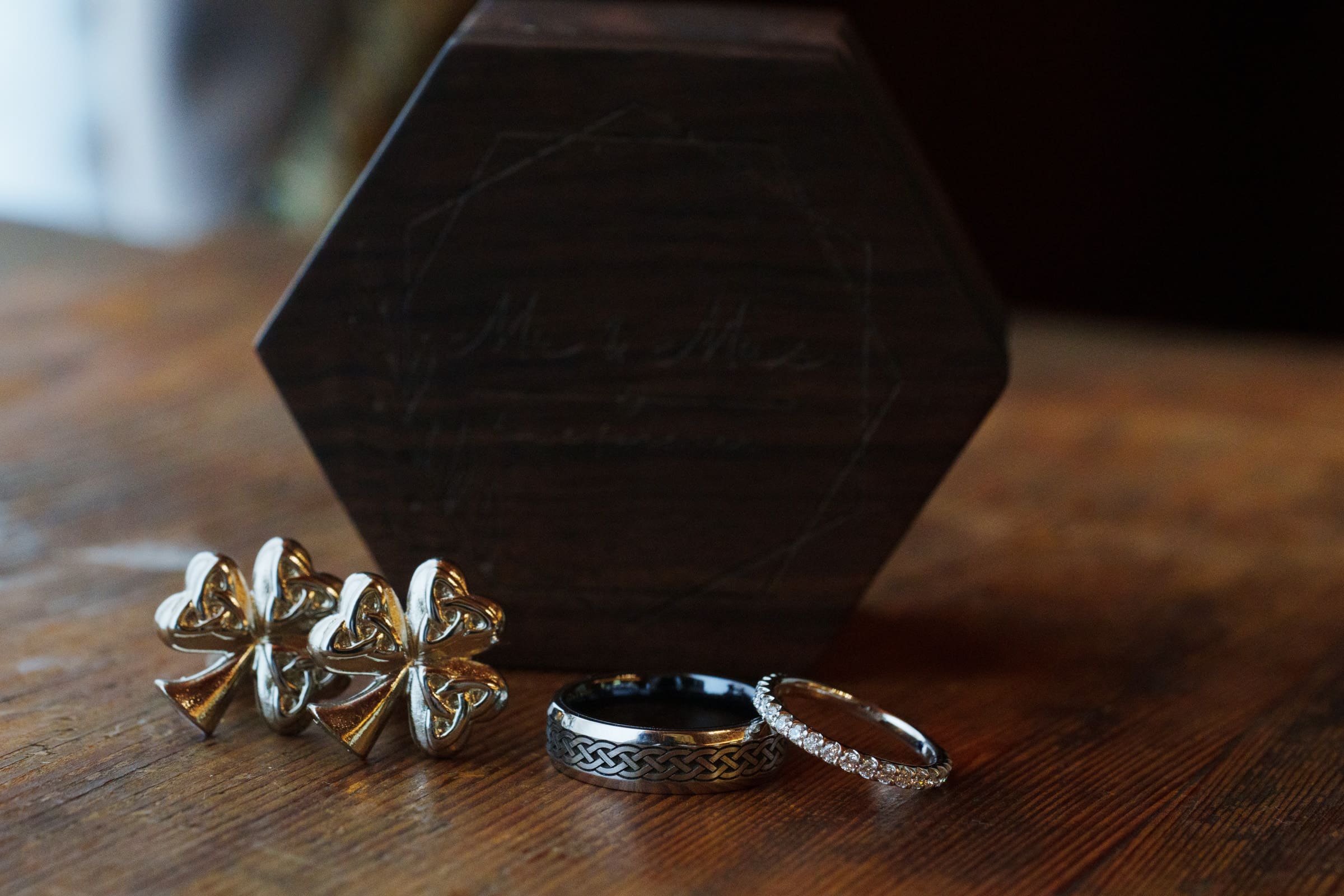 Close-up of a wooden hexagonal box with Mr. & Mrs. engraved on it, reminiscent of a Rose Hill Estate wedding. Placed on a wooden surface, two cufflinks are accompanied by a black and silver patterned ring and a silver band adorned with small gemstones.