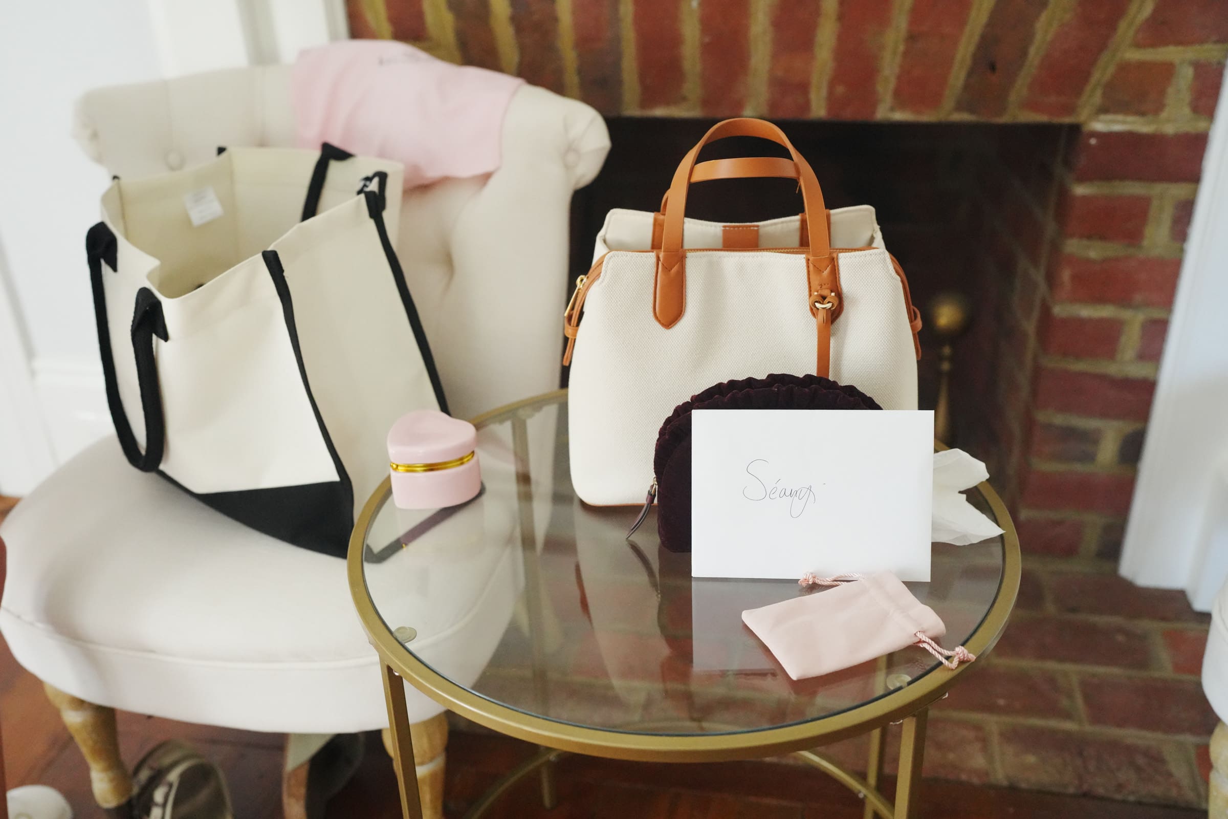 A cozy setting reminiscent of a Rose Hill Estate wedding, with a brick fireplace in the background. Two stylish bags and small pouches rest on a glass table and chair. A card with Savvy written on it is placed on the table, alongside a small round box, creating an inviting scene.