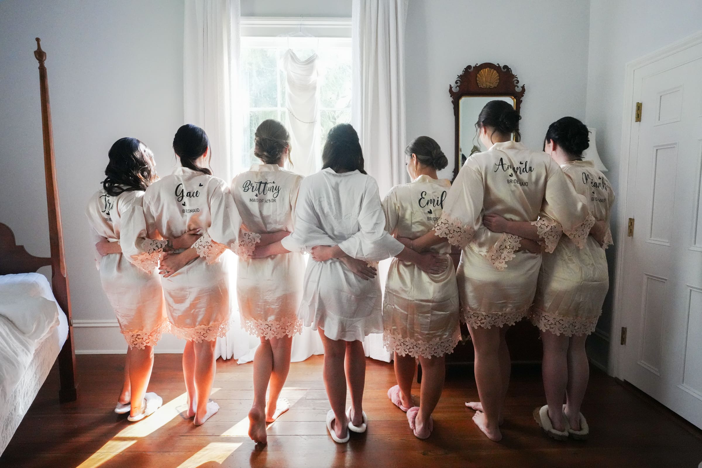 A group of seven women wearing matching cream-colored robes with names on their backs stand in a line, arms linked, facing a window with a hanging wedding dress. In the Rose Hill Estate wedding venue, the room boasts wooden floors and a vintage mirror, exuding timeless elegance.