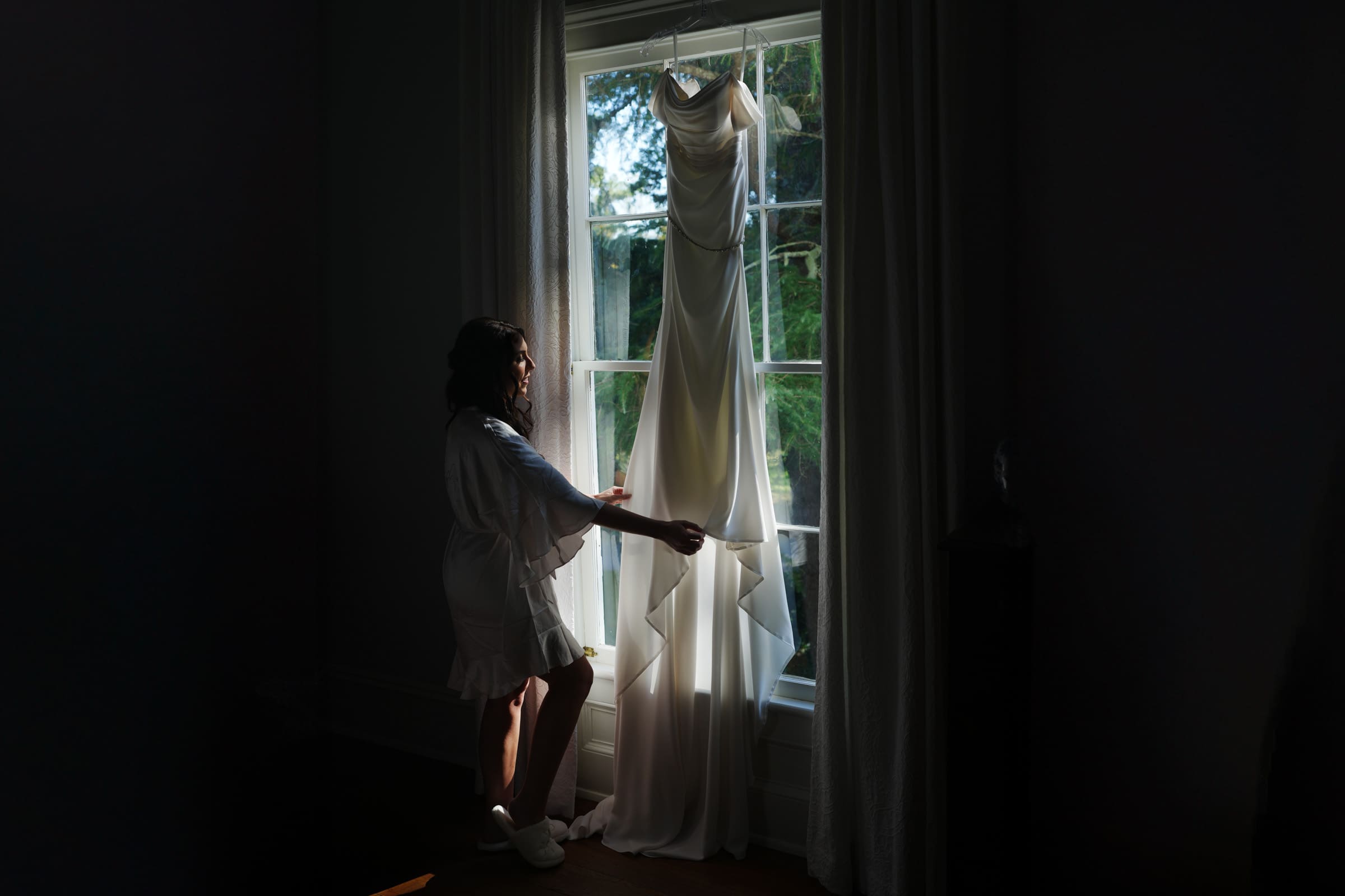 A woman in a robe stands by a large window at Rose Hill Estate, gently touching a flowing white dress hanging there. Sunlight streams through the window, illuminating both the dress and the lush greenery outside, setting the perfect scene for a wedding.