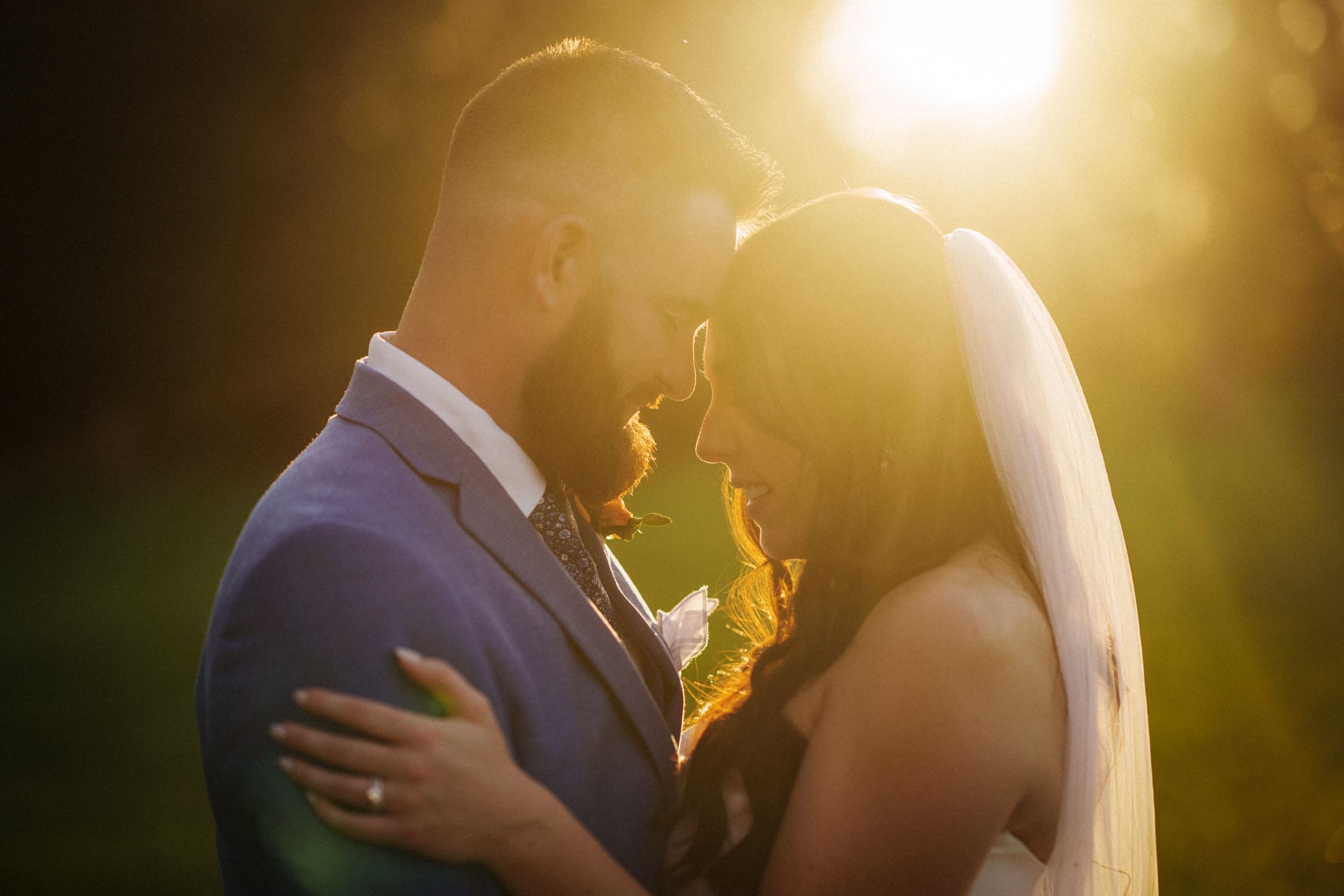 A bride and groom embrace during sunset at Rose Hill Estate, bathed in golden light. The groom wears a blue suit with a boutonniere, while the bride, in a white gown and veil, touches his tie. They stand outdoors, surrounded by a soft, warm glow.