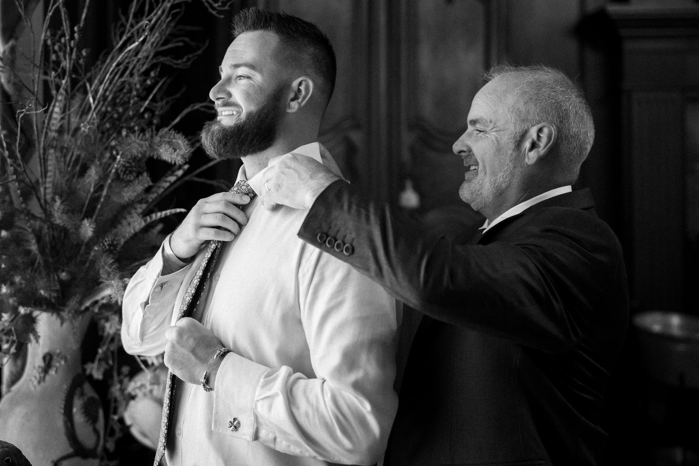 A man helps another adjust his tie, both smiling in formal attire at a Rose Hill Estate wedding. The setting is indoors with decorative plants in the background. The image is elegantly captured in black and white.